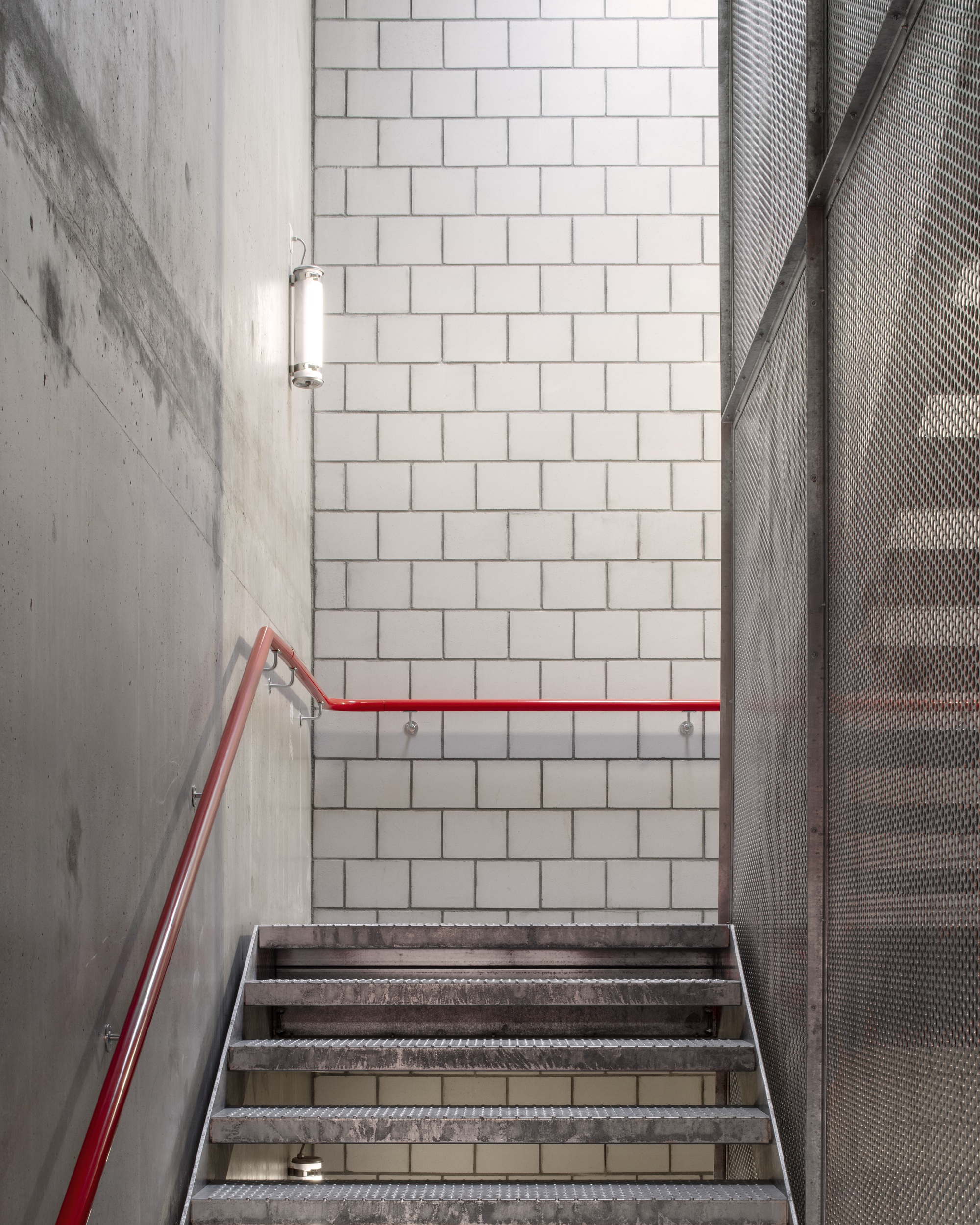 Concrete stairwell with red handrail, white tile wall and perforated metal screen panel