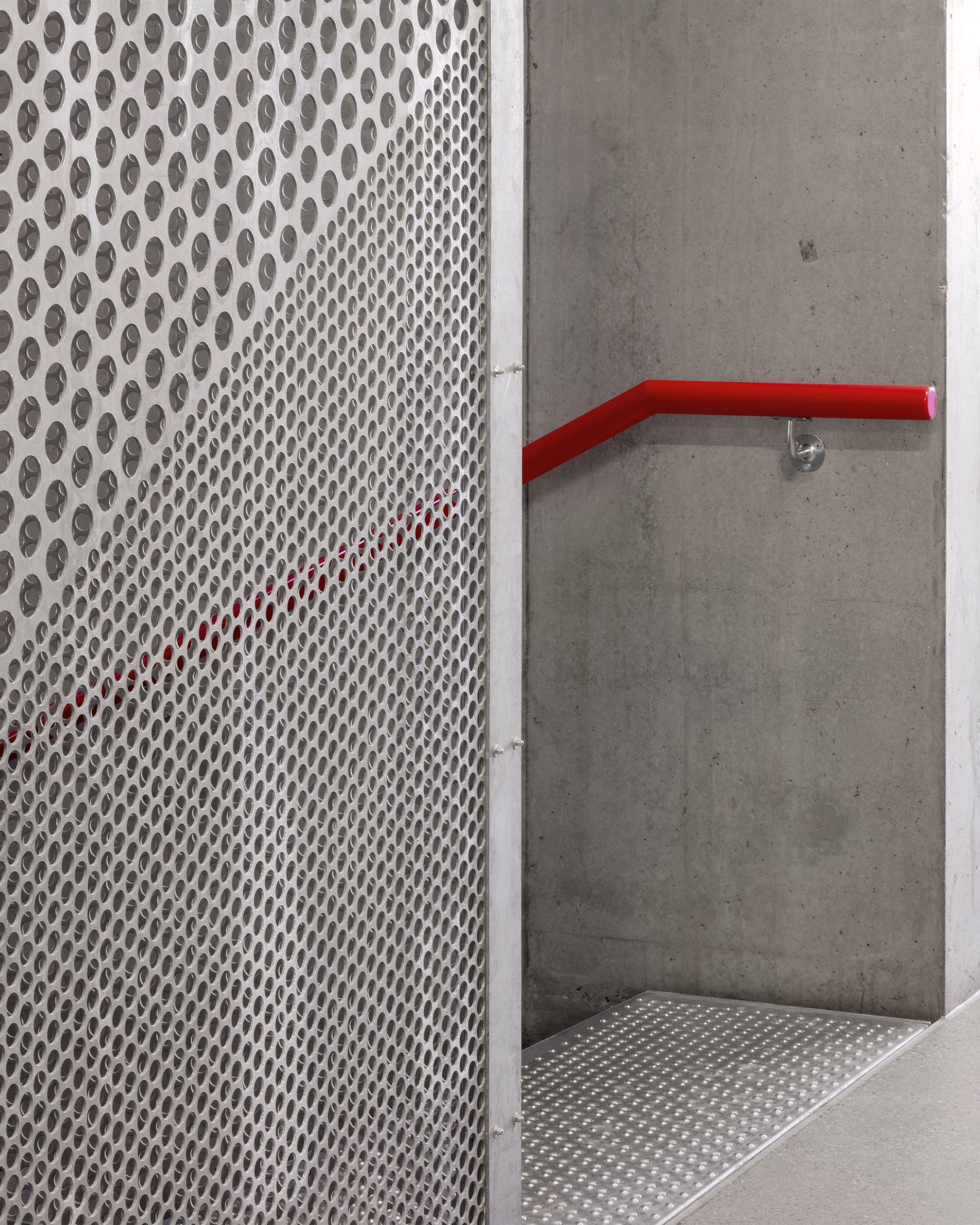 Perforated metal screen wall meeting a concrete wall with a red painted handrail at the landing