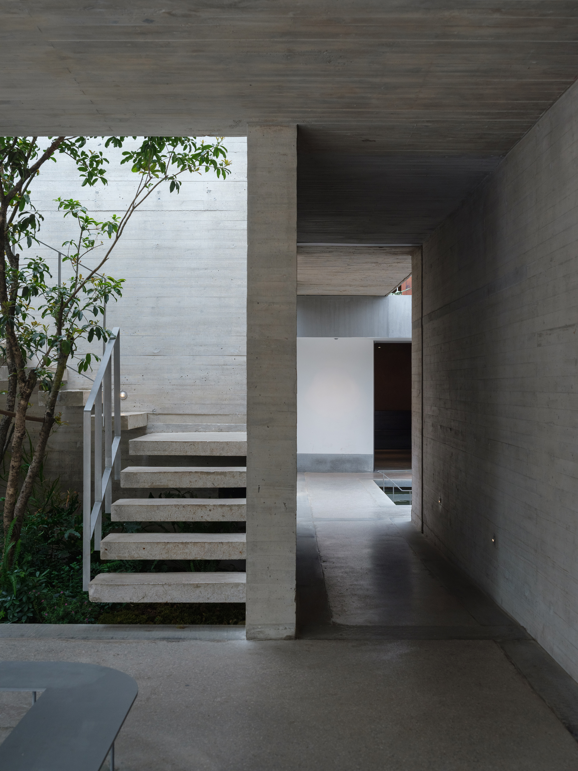 Concrete corridor with cantilevered staircase and tree in adjacent courtyard under overcast sky