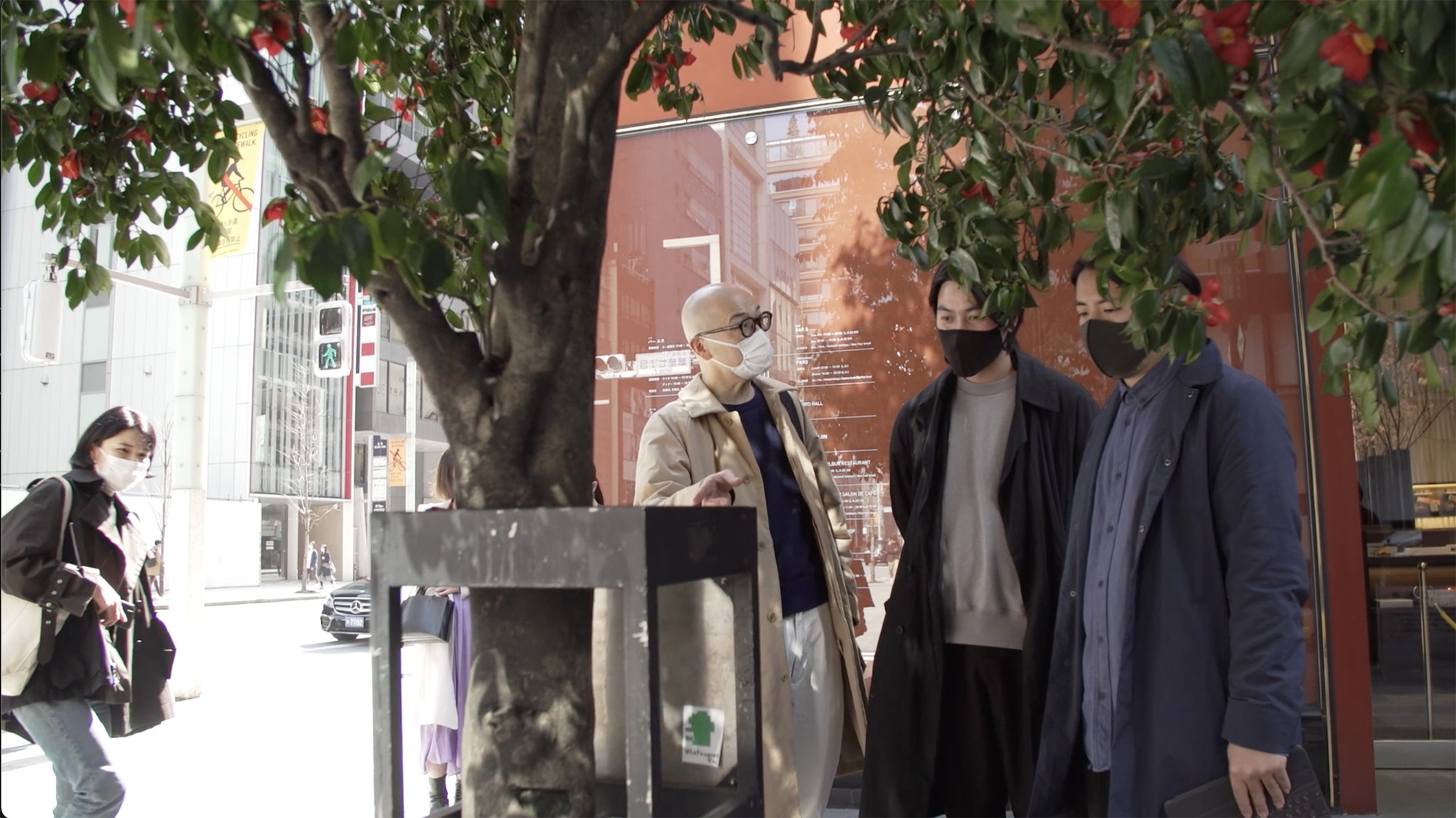 Four people gathered beneath a tree outside a glass-fronted exhibition space in daylight