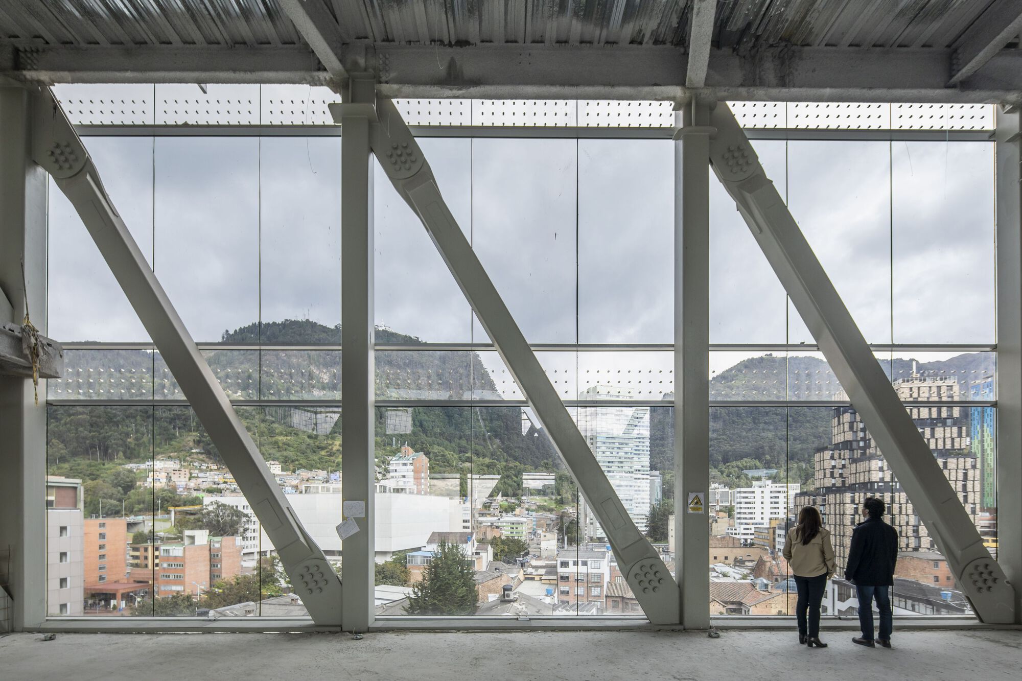 Full-height window wall with diagonal white structural bracing as two people overlook the surrounding cityscape