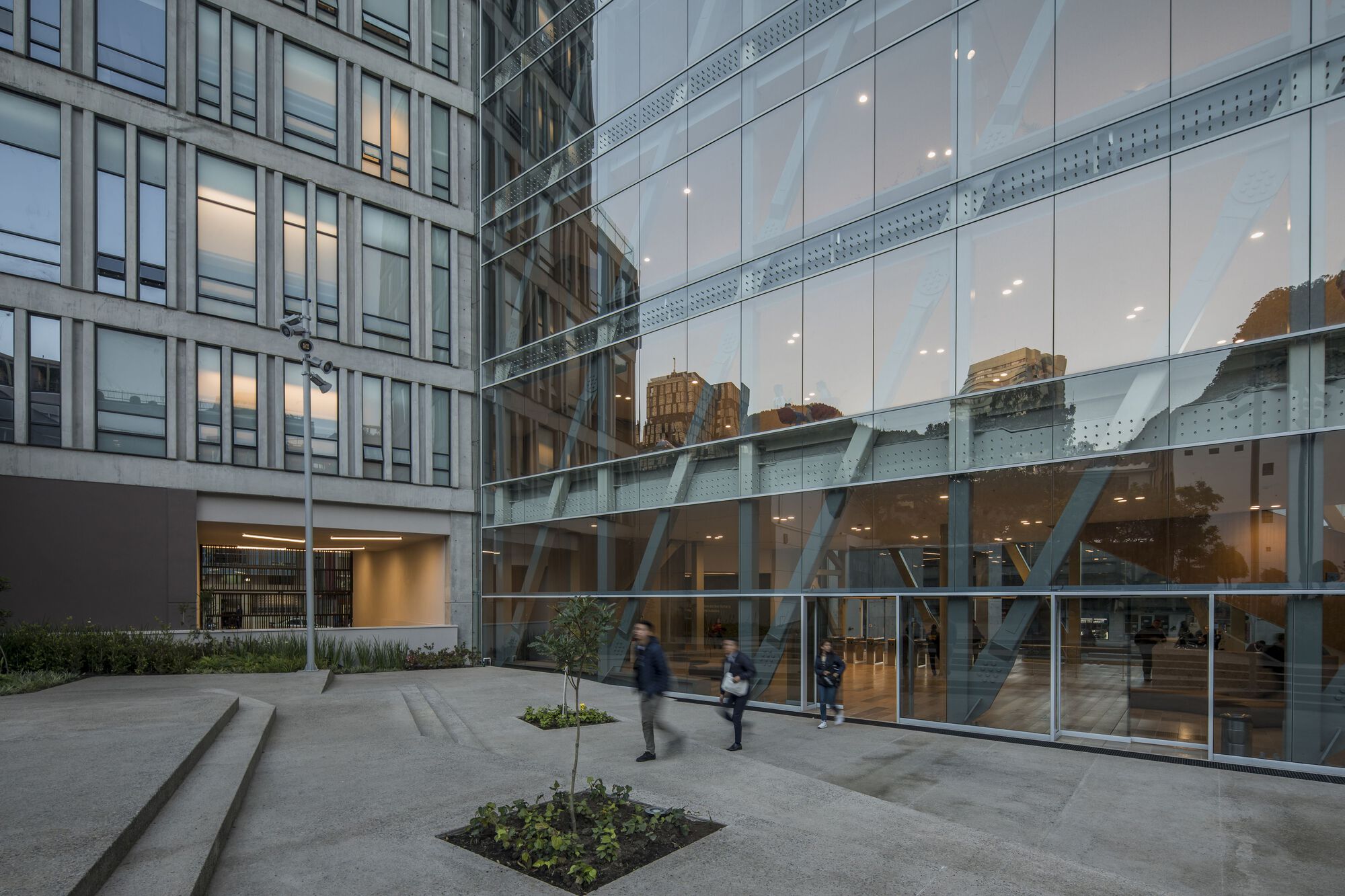 Evening courtyard view showing the angled steel braces behind the glass ground floor facade with people passing