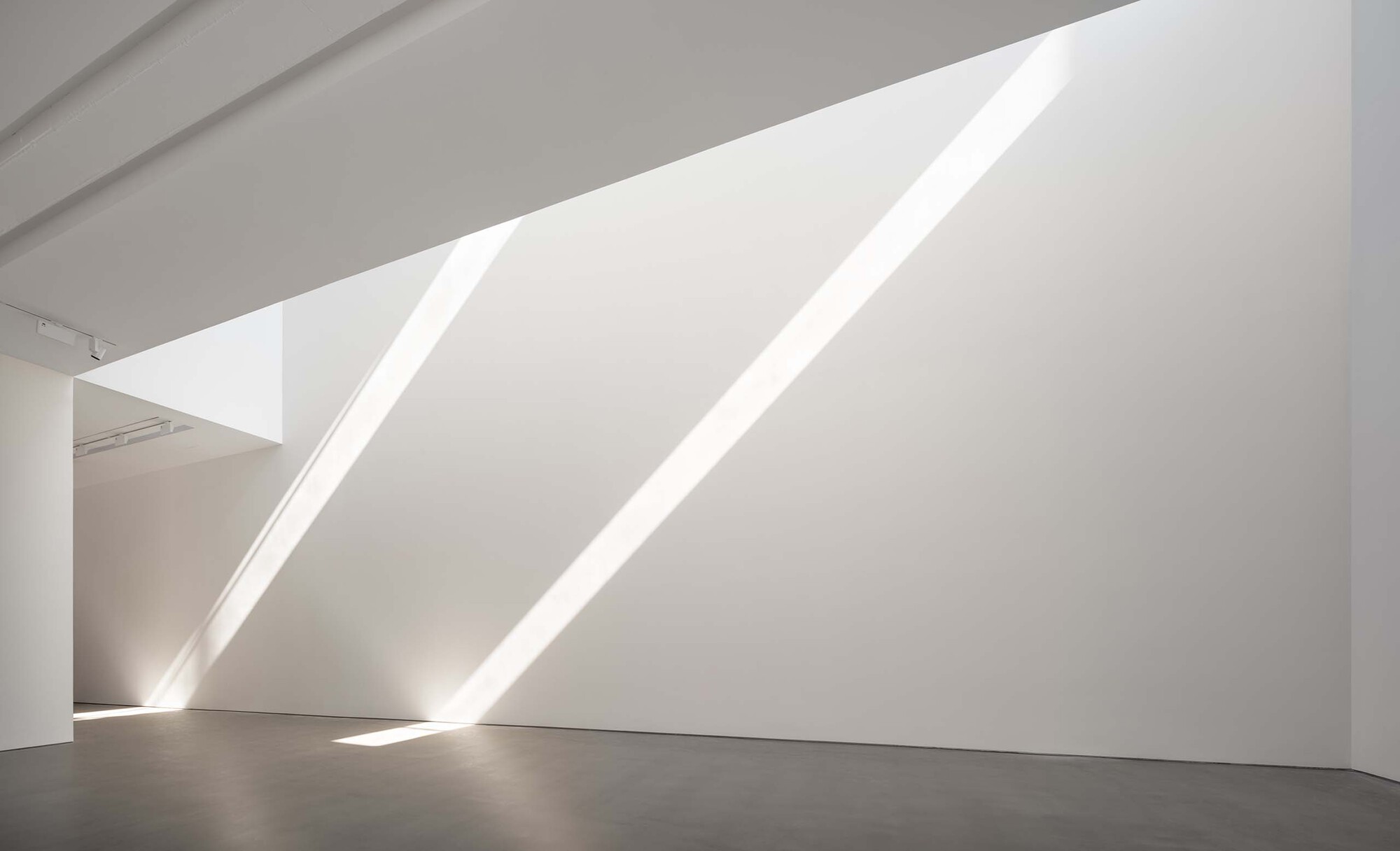 White gallery wall with diagonal skylight slots casting sharp shadows across the polished floor