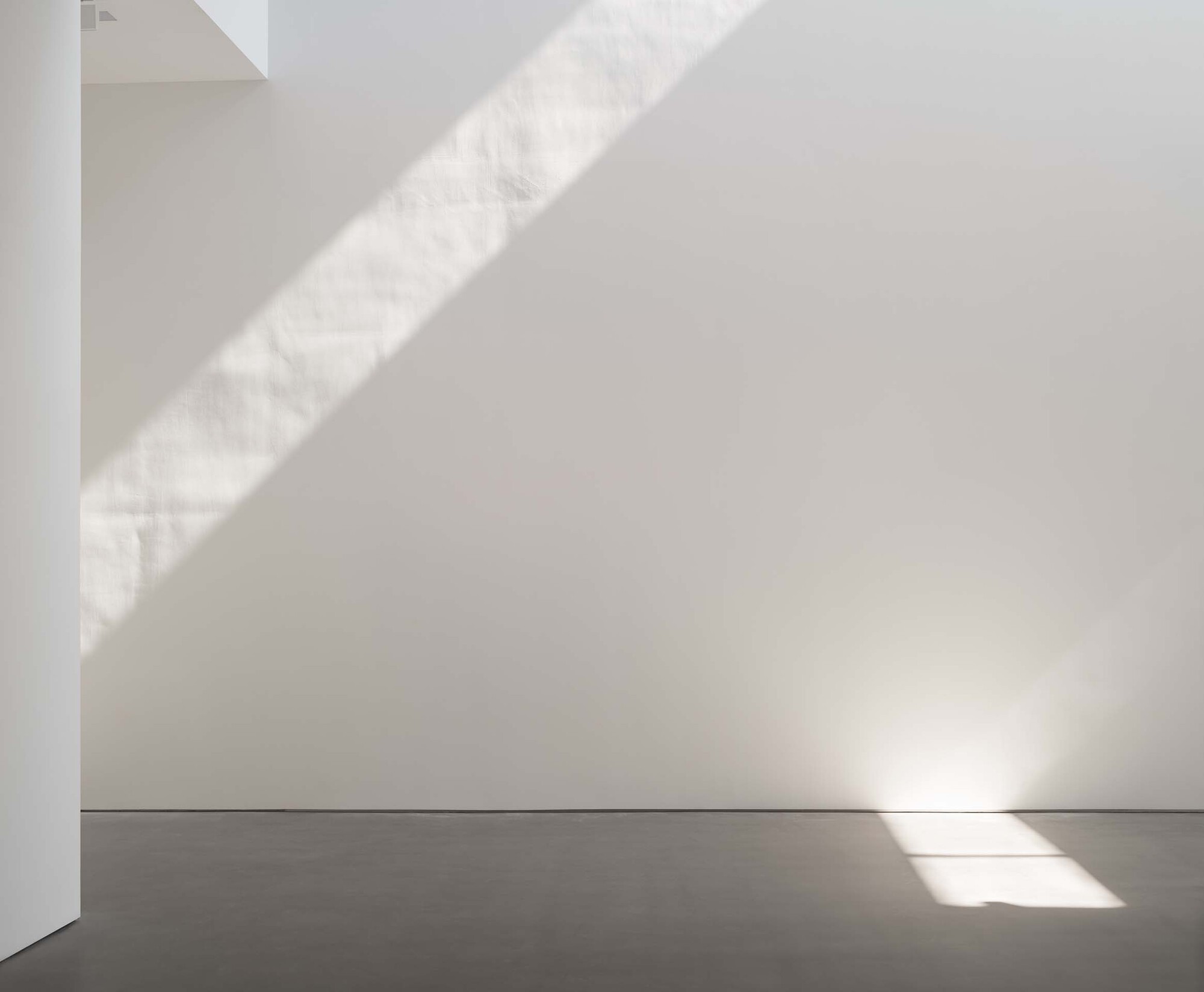 White interior corner with diagonal shadows cast by unseen skylight above grey concrete floor