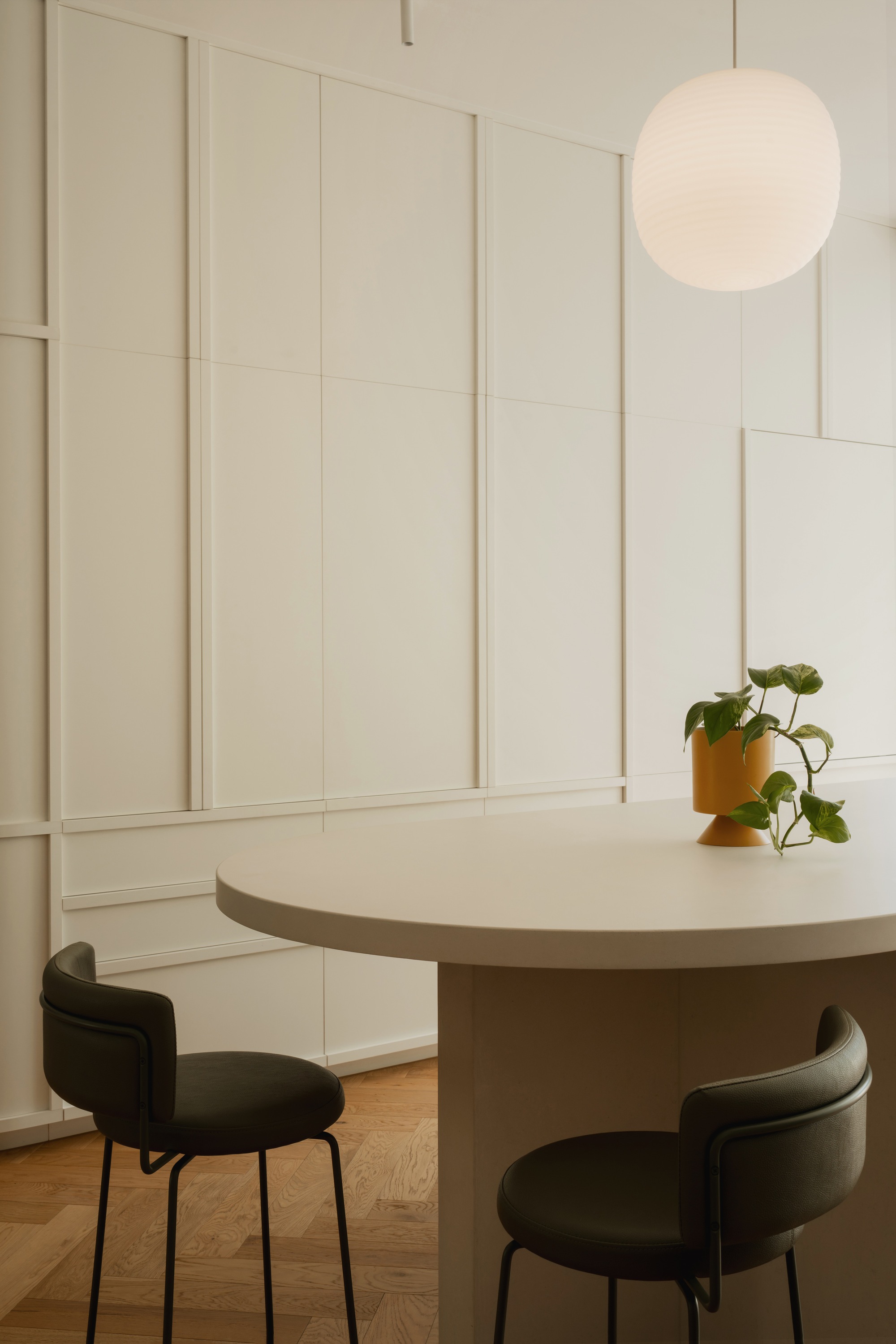 Round dining table with dark upholstered chairs and potted plant under spherical pendant at dusk