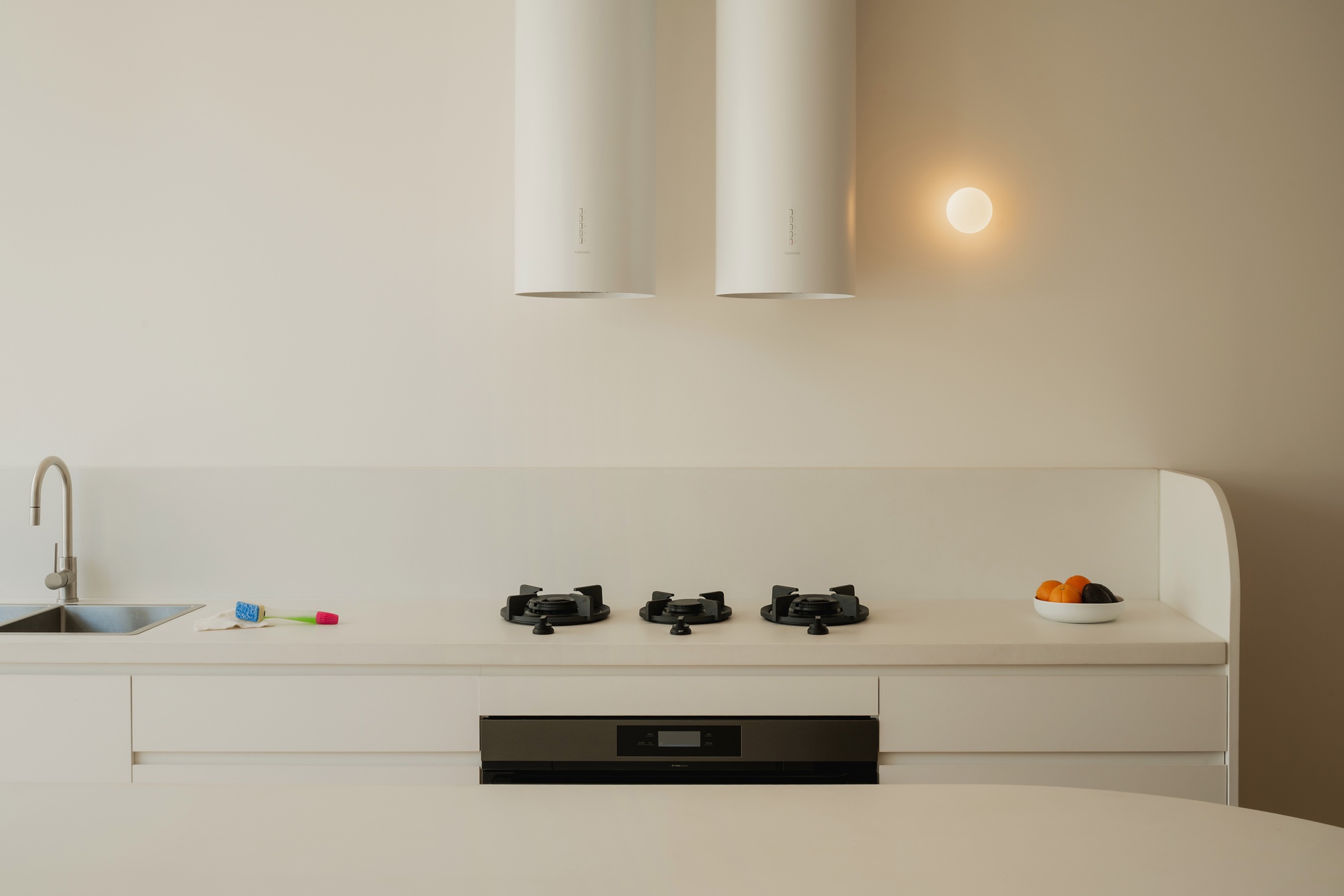 Minimalist kitchen with white countertop, three gas burners, and cylindrical range hood under warm evening light