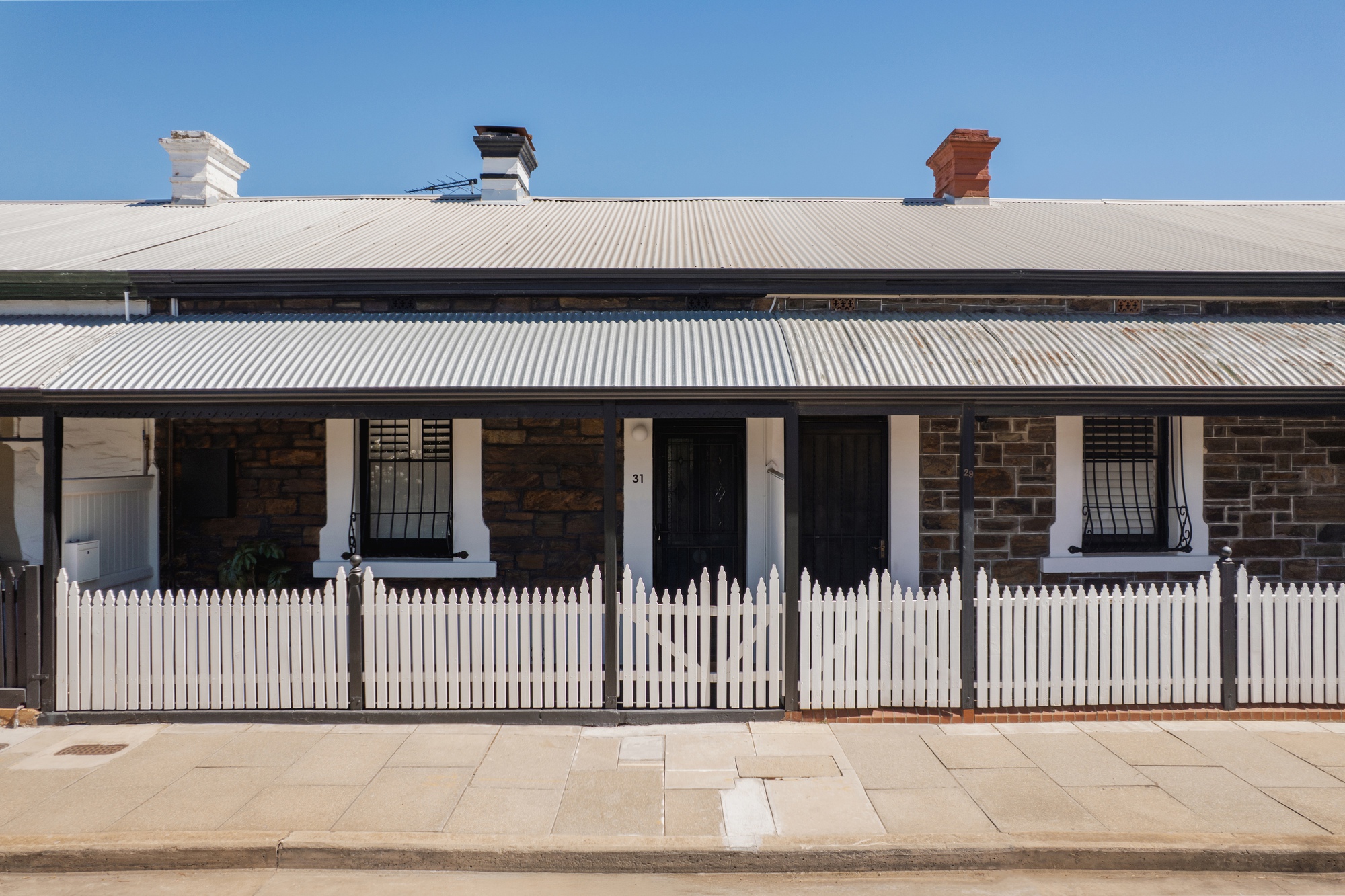 Street view of a stone cottage with corrugated metal roof, white picket fence and verandah under clear blue sky