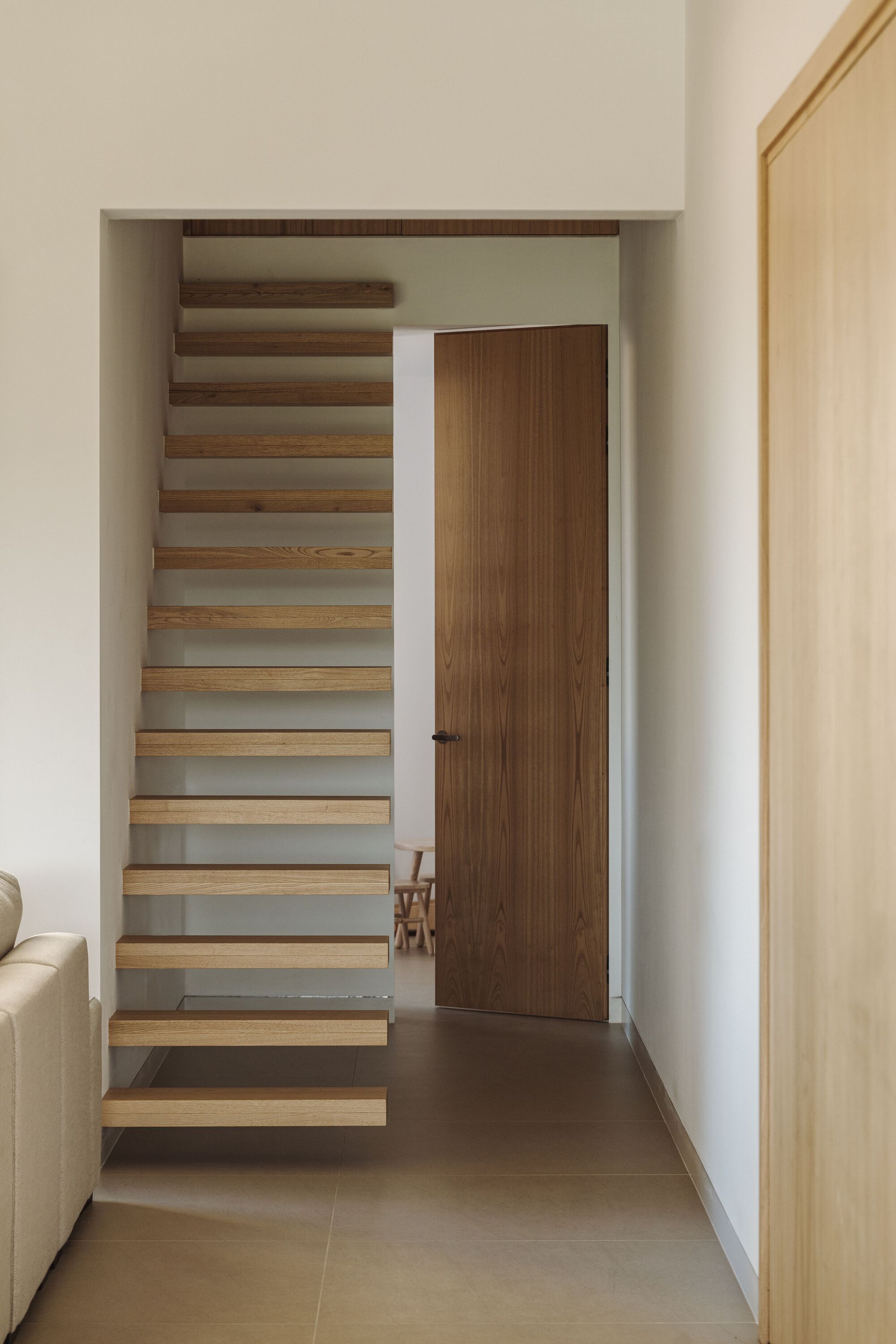 Floating timber staircase with open risers rising beside a closed wooden door in a white hallway