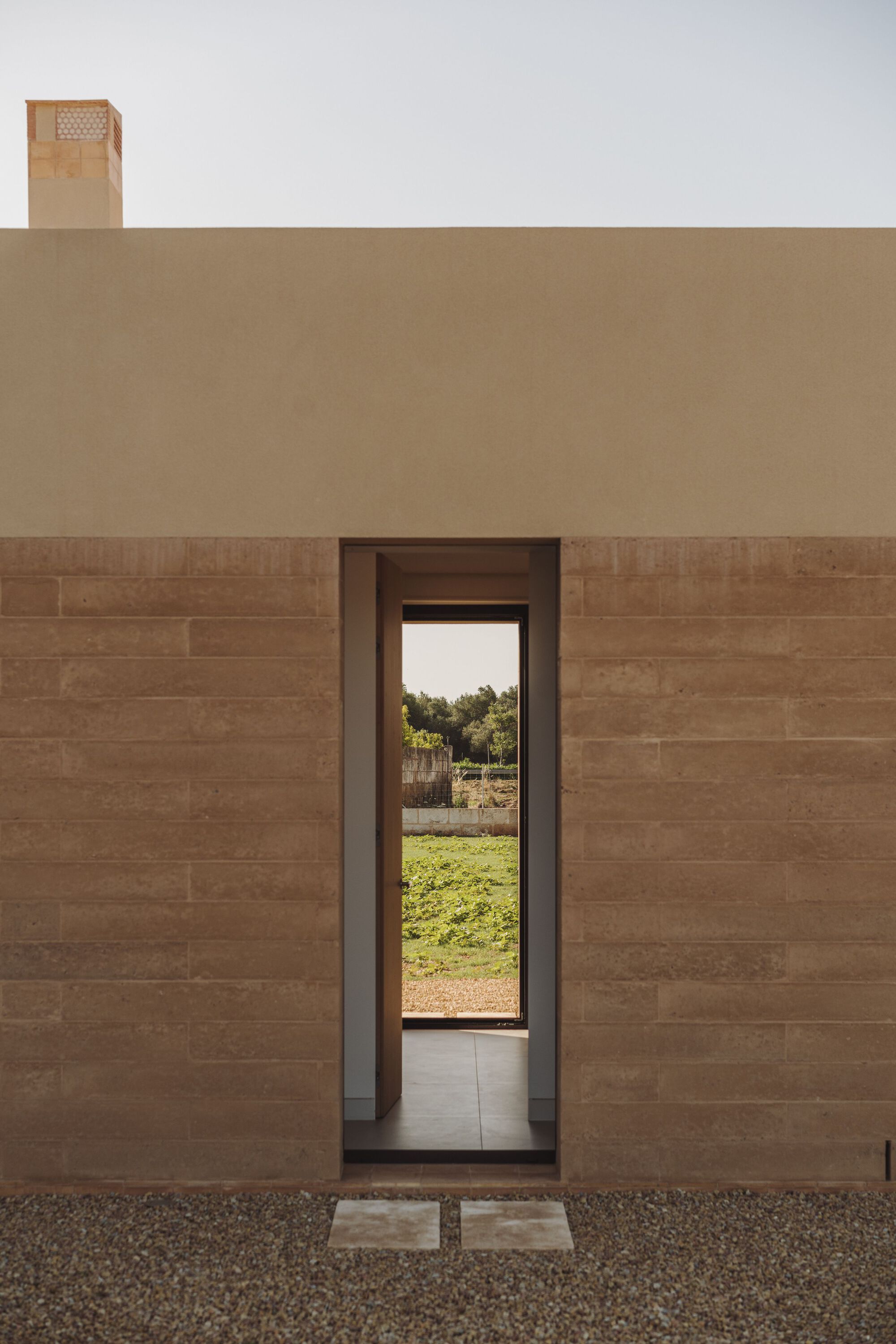 Tall narrow doorway framing view of gravel courtyard and green lawn beyond rammed earth facade