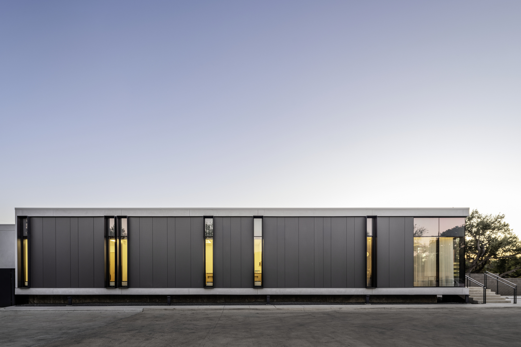 Street-facing facade with dark metal panels and vertical slot windows illuminated from within at dusk
