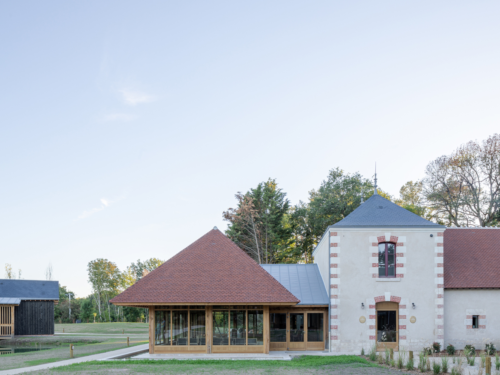Glazed pavilion with pyramidal tile roof next to a rendered tower with brick quoins at dusk