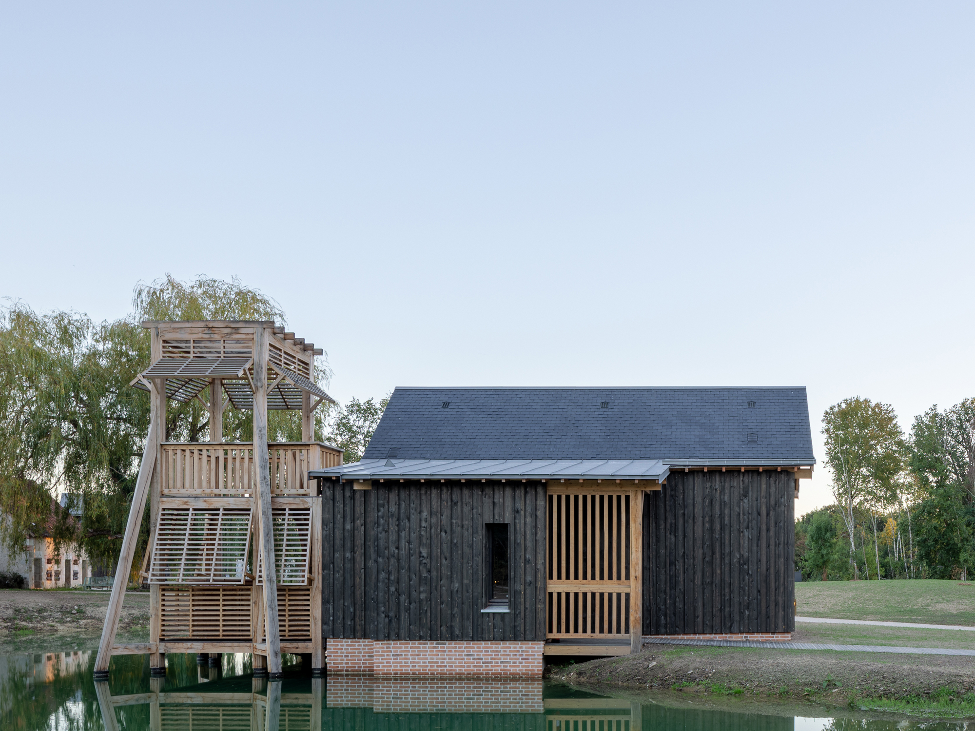 Charred timber volume on a brick plinth beside a timber observation tower reflecting in a pond