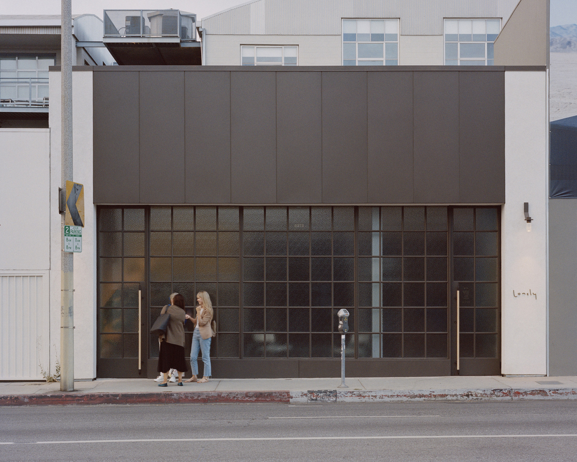 Street facade with gridded glass storefront and dark metal panel upper volume as two pedestrians pause at the entrance