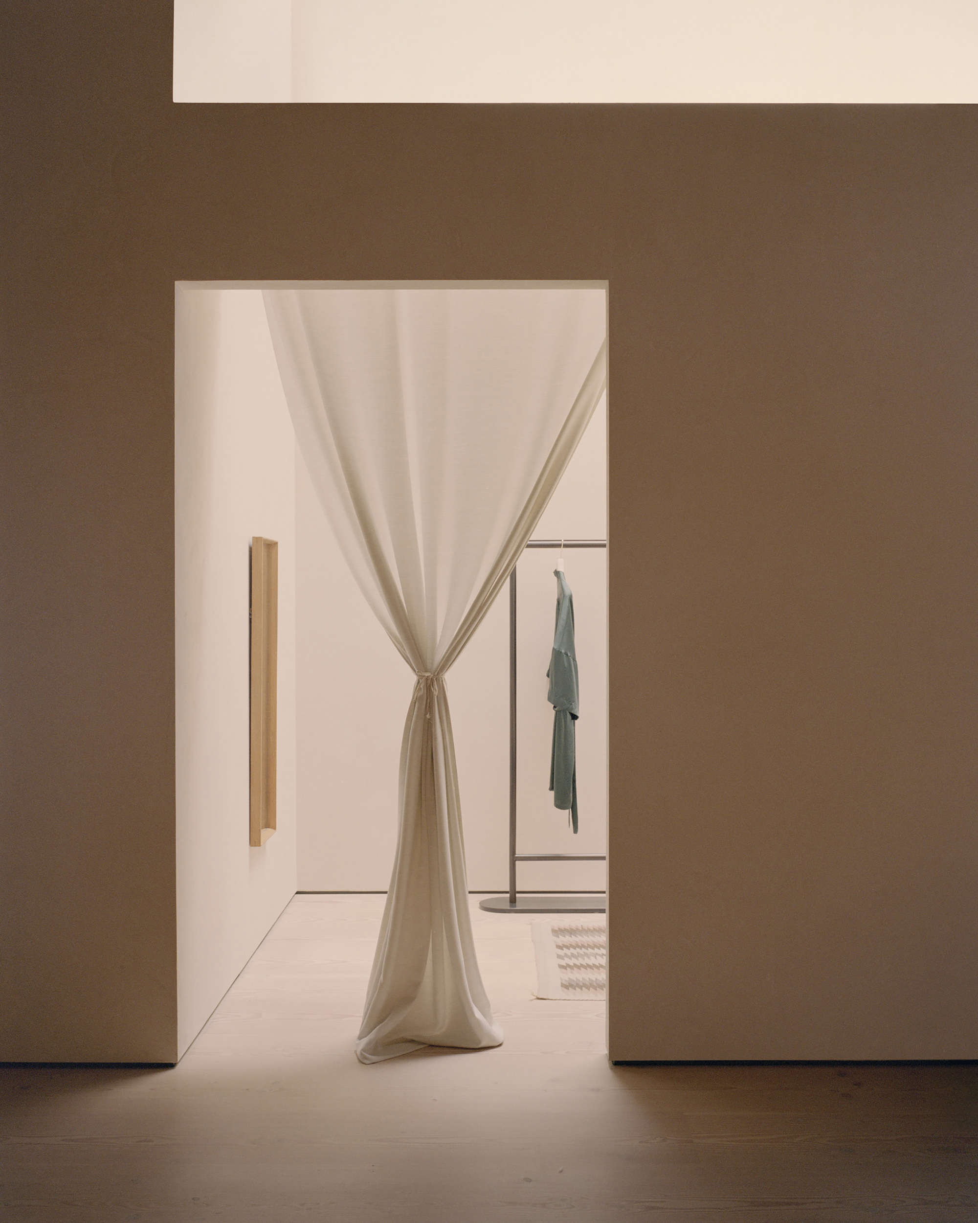Doorway framed in warm brown walls with draped linen curtain tied back, revealing clothing rack beyond