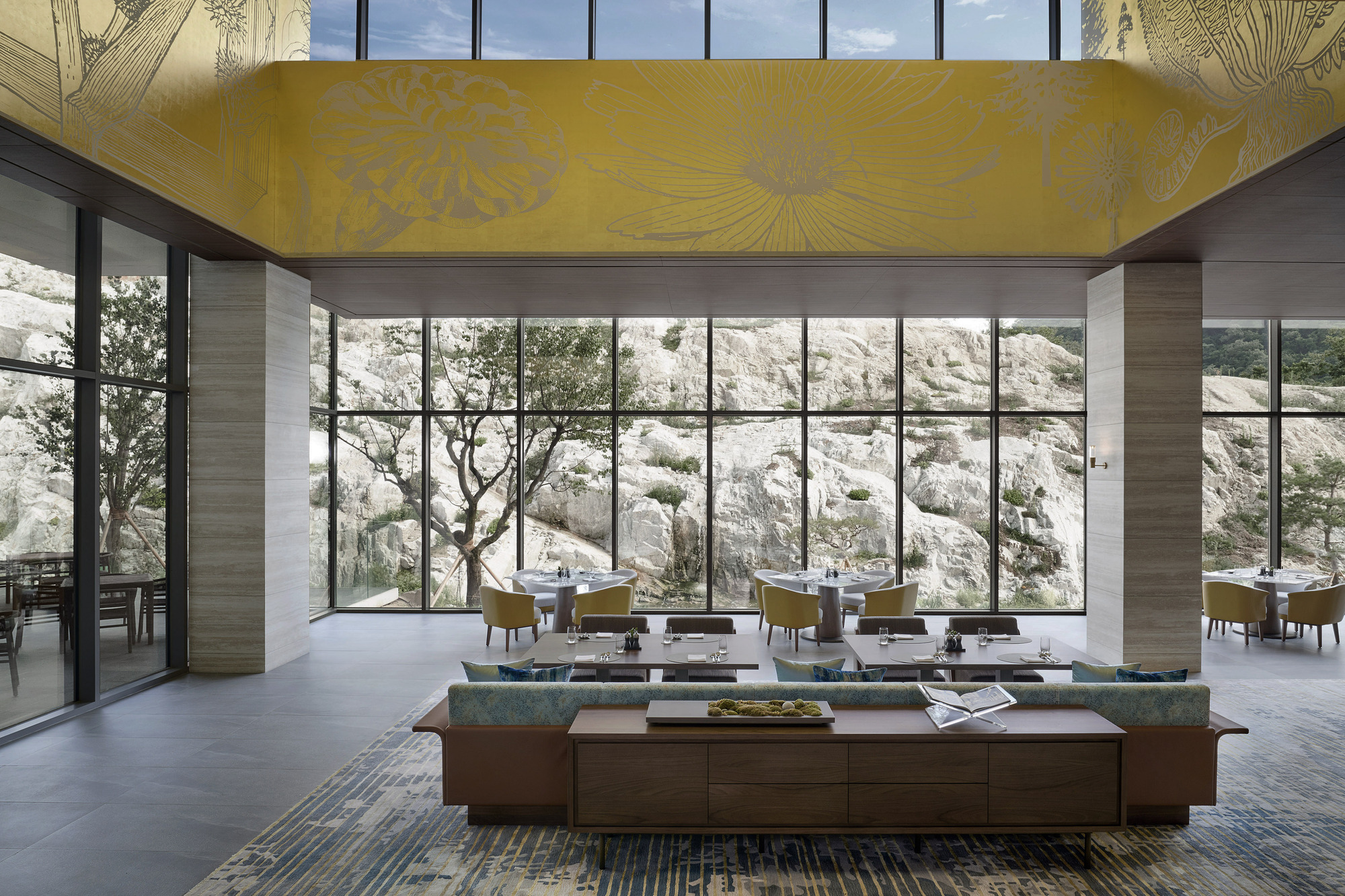 Double-height lounge space with yellow floral ceiling panel and floor-to-ceiling glass overlooking the limestone cliff