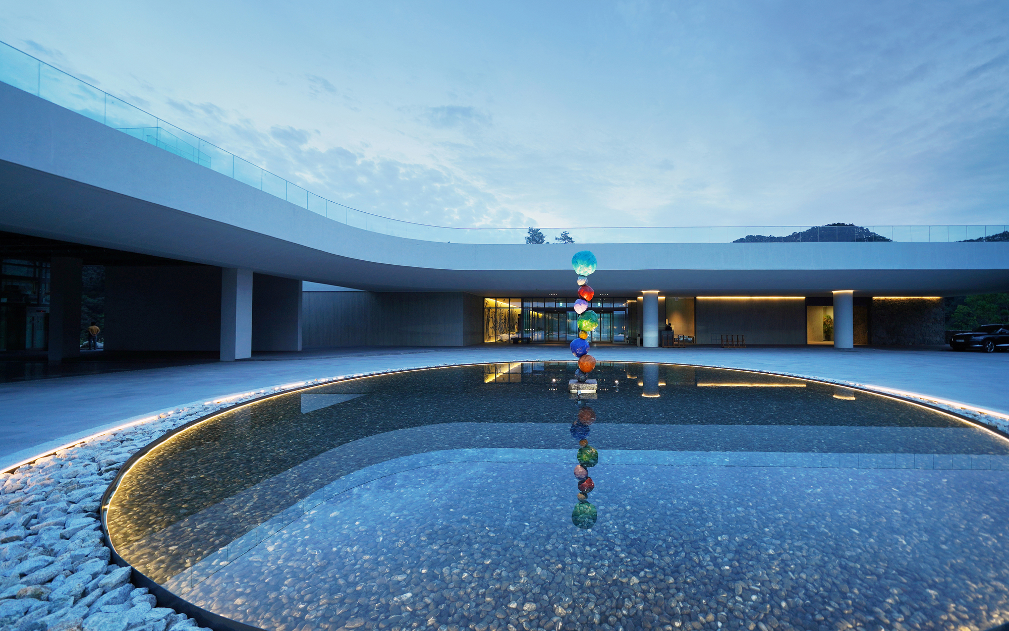 Curved courtyard pool with sculptural installation reflecting the glazed colonnade and mountains beyond at twilight