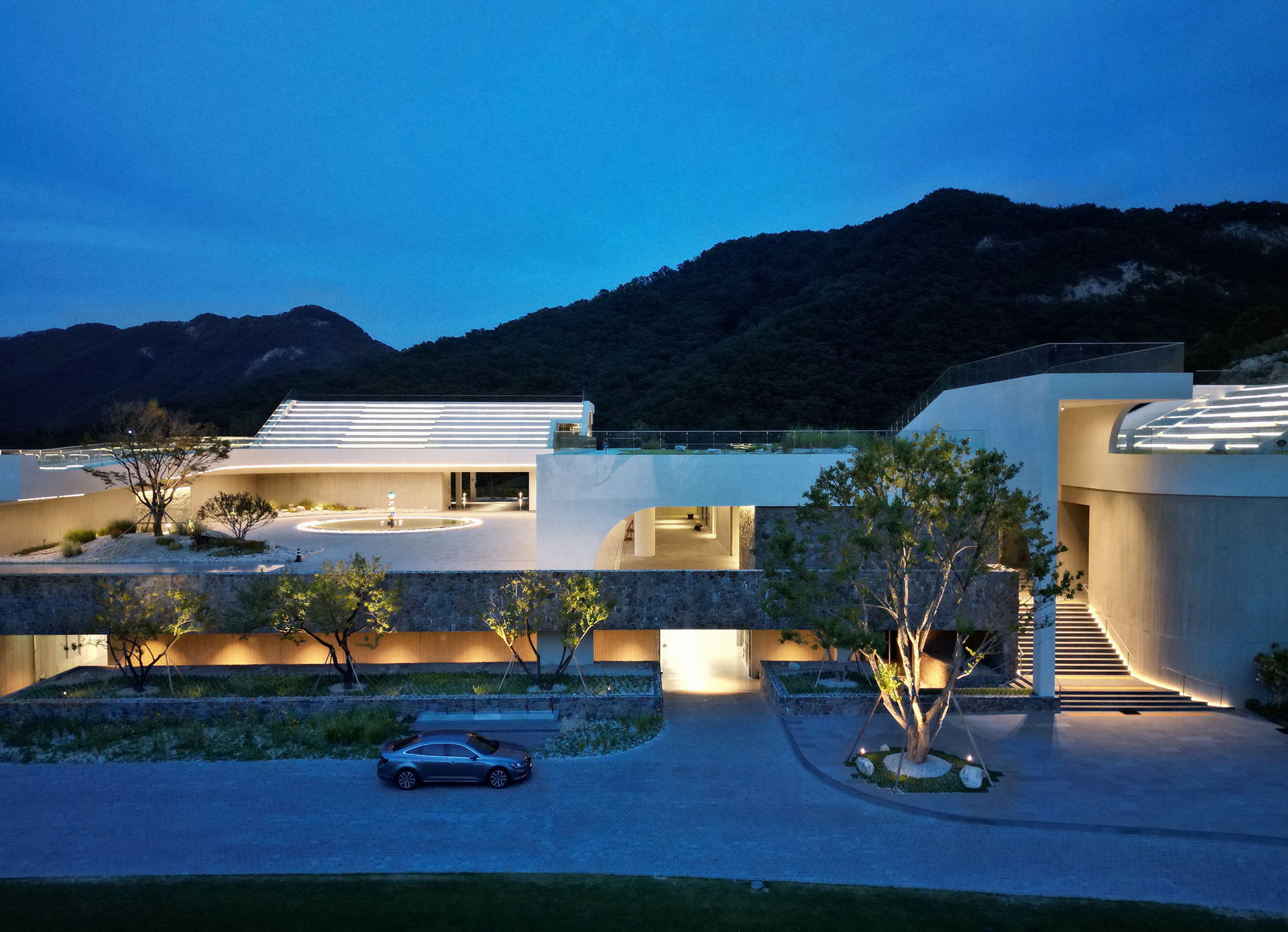 Night view of the white horizontal volumes with ribbed roofs and illuminated openings beneath the dark mountains