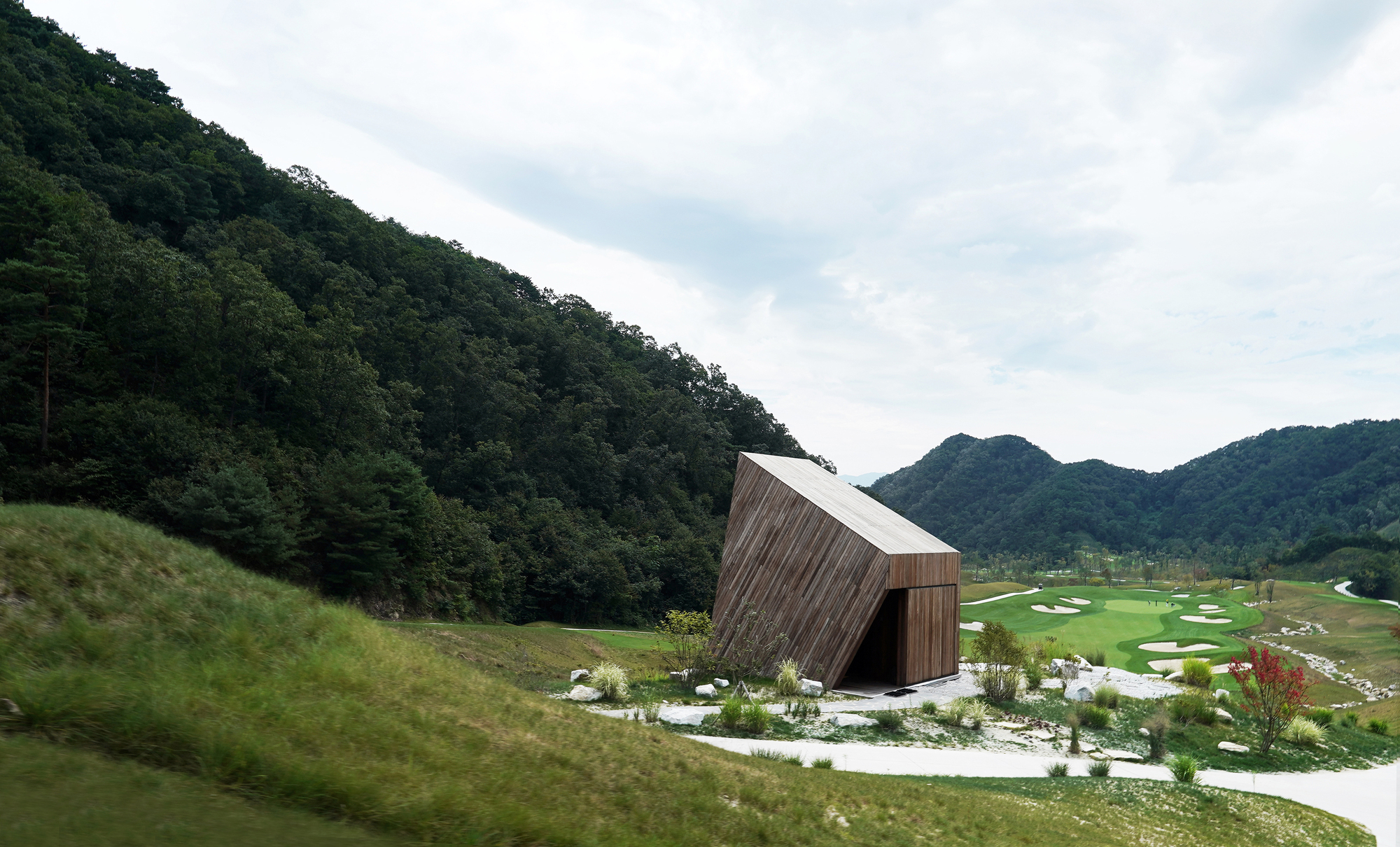 Timber-clad pavilion with angled roof set on a hillside overlooking the golf course in daylight
