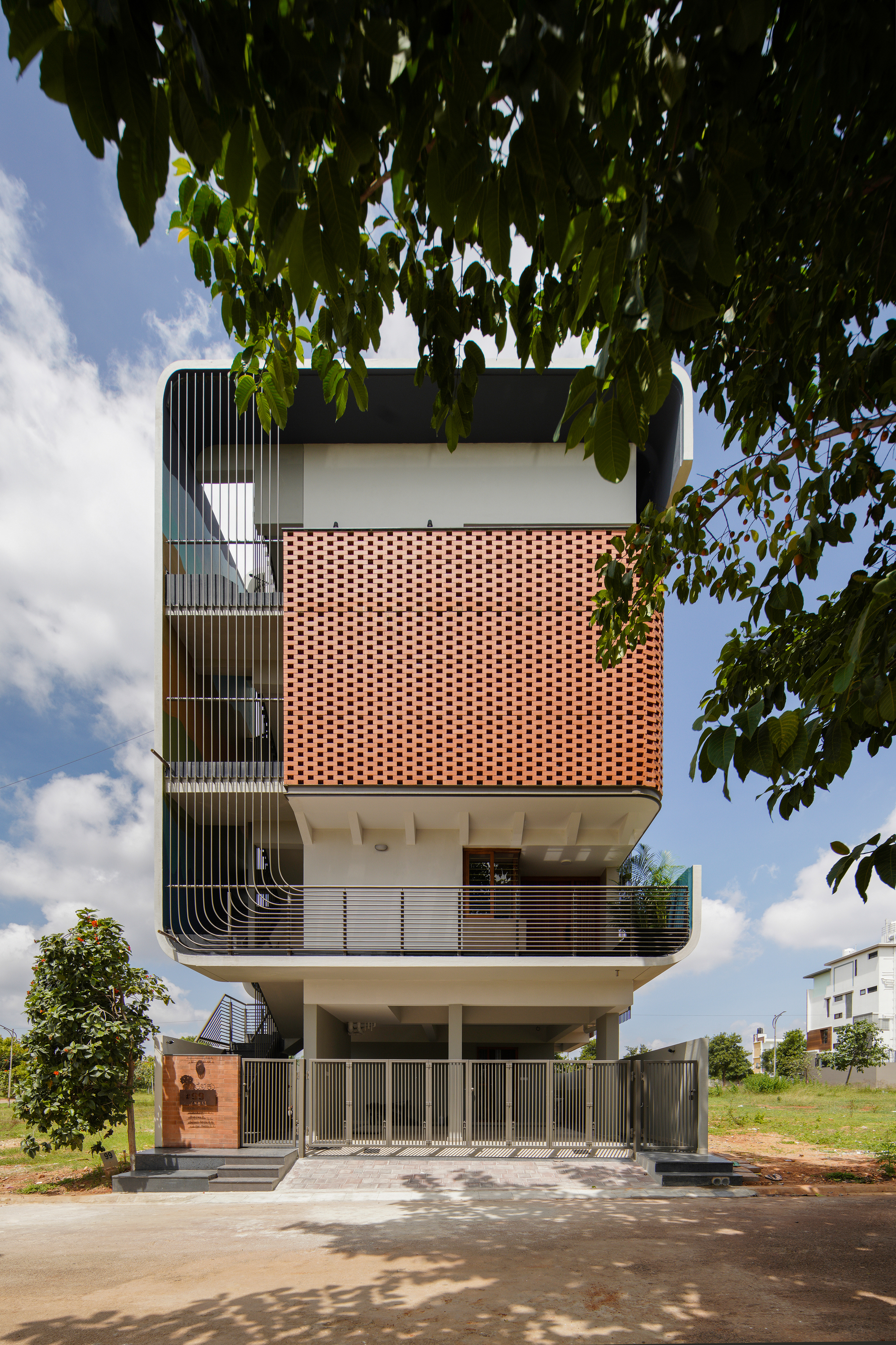 Front elevation showing vertical louver panels on the left and a perforated terracotta volume under afternoon clouds