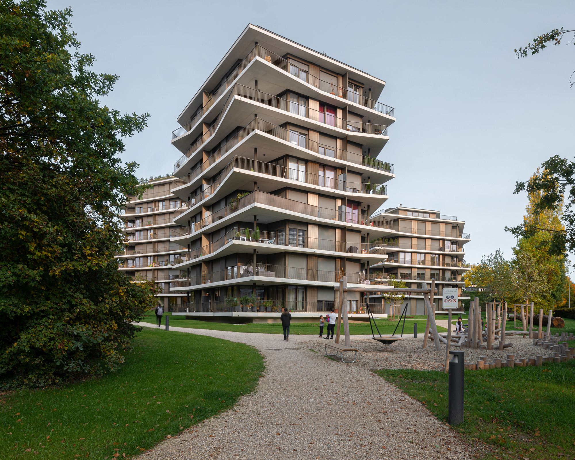 Stepped residential tower with deep concrete balconies and timber screens seen from a park pathway