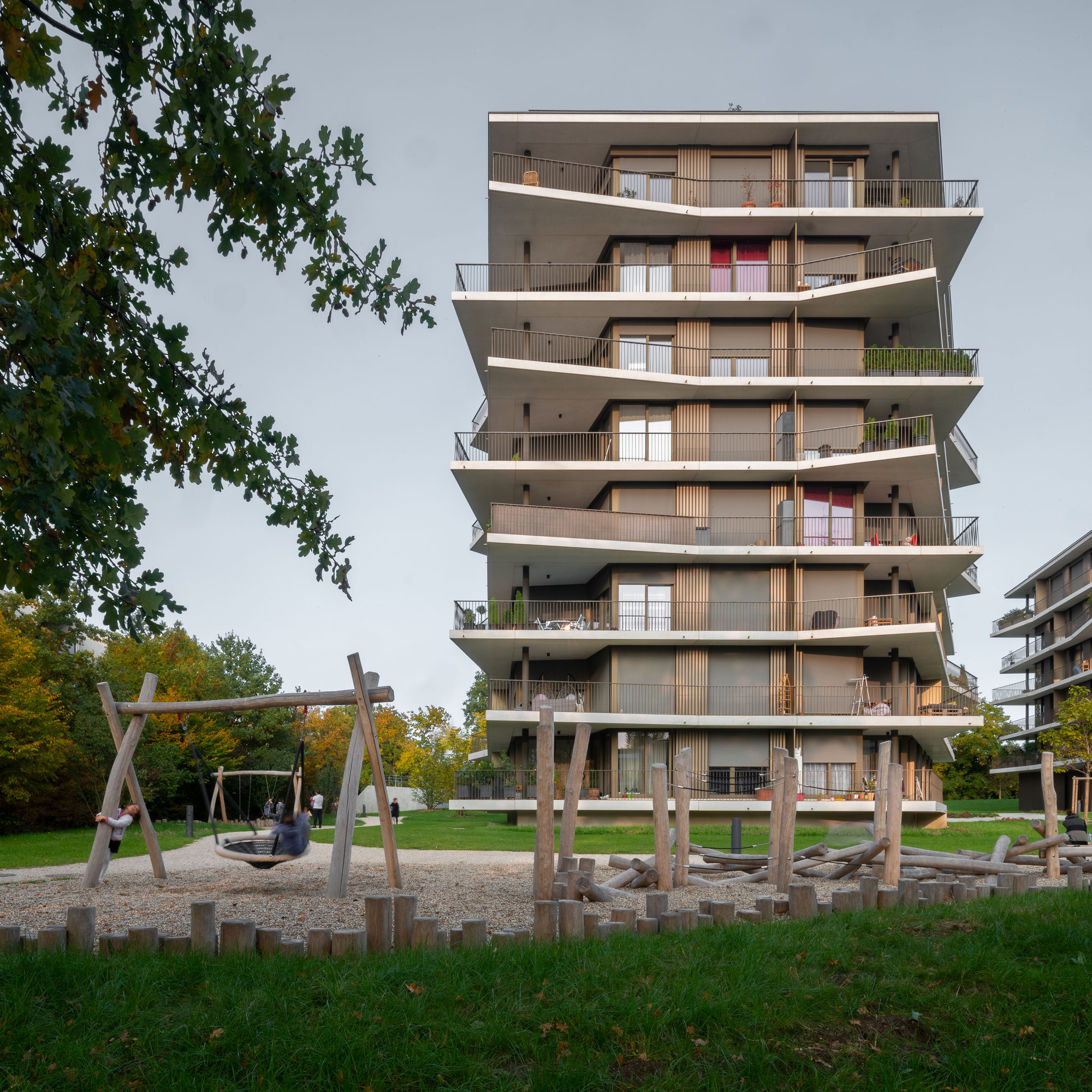 Tower facade with cantilevered concrete floor plates and timber balustrades above a playground with wooden logs