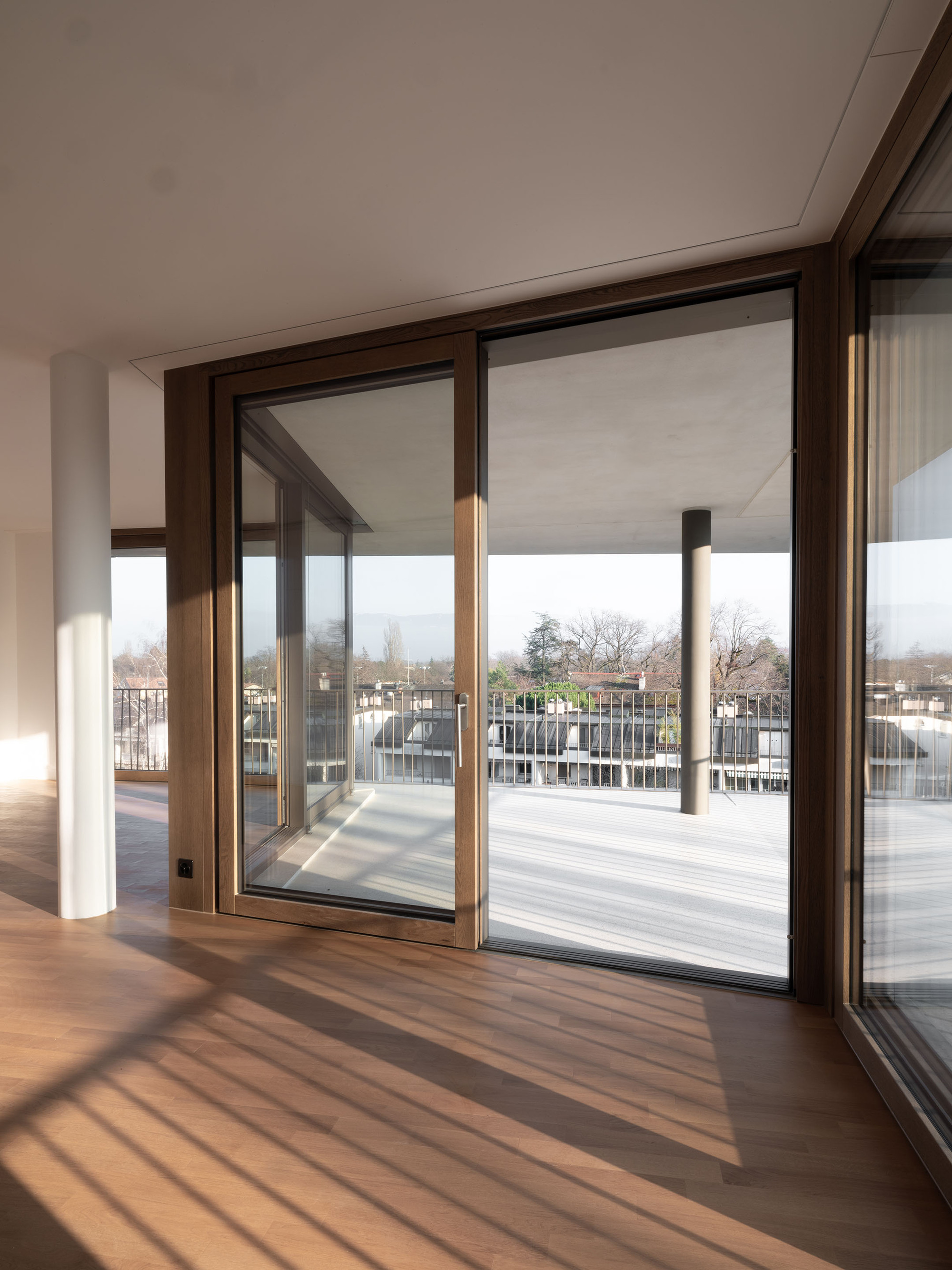 Interior view through bronze-framed sliding doors opening to a terrace with strong diagonal shadows on timber flooring