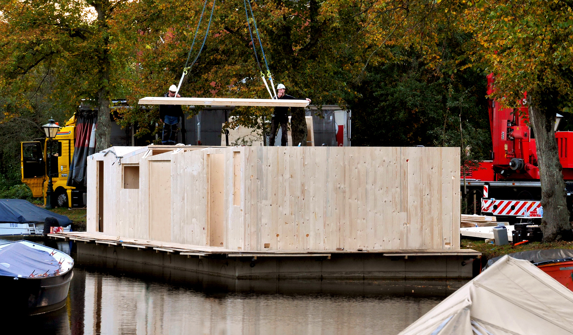 Prefabricated timber-clad floating structure being lifted by crane during assembly on the water