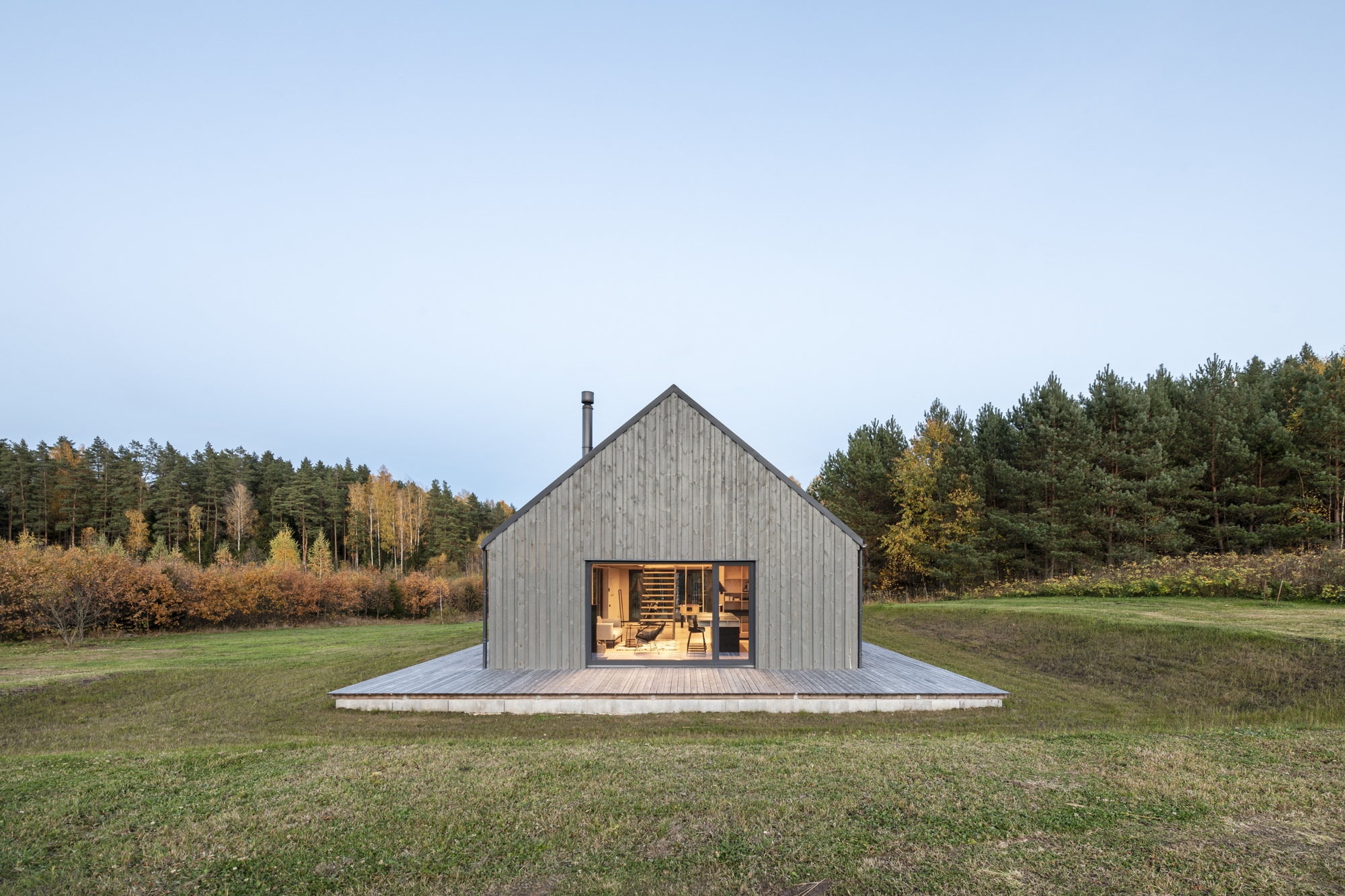 Gabled end facade with full-height glazing and timber deck surrounded by meadow and forest