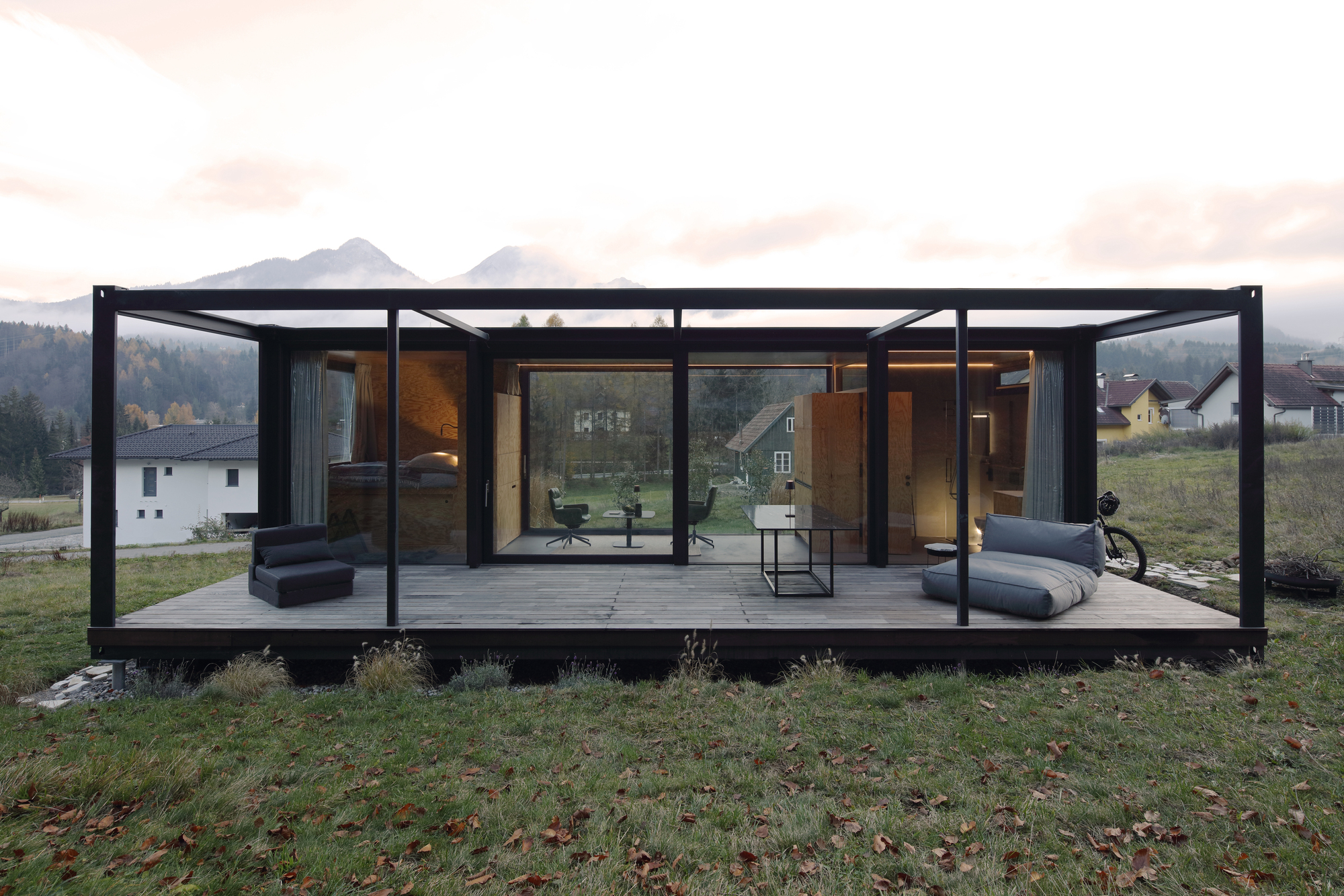 Glass-walled pavilion with black steel frame and covered terrace overlooking a mountain valley at dusk