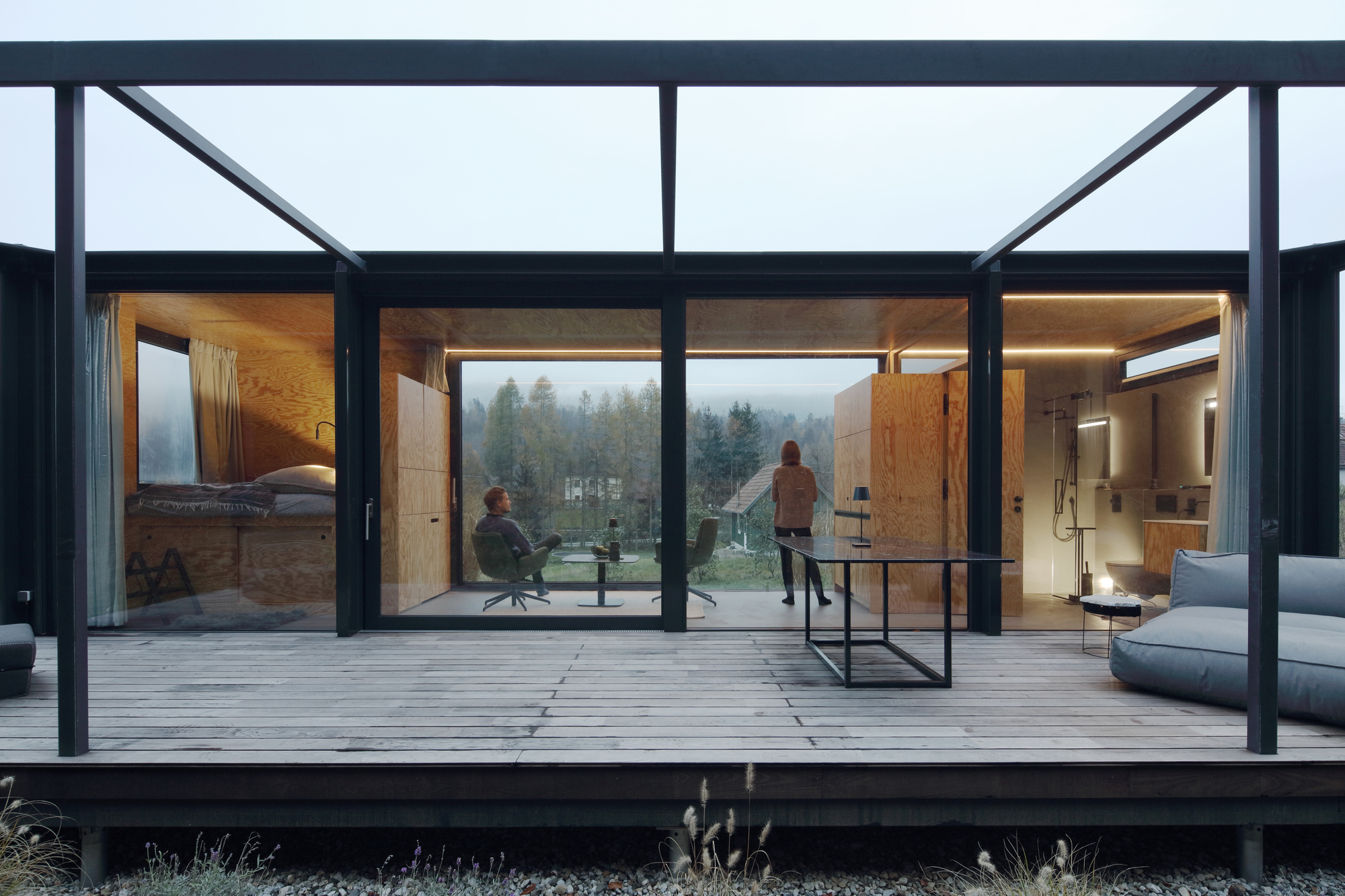 Timber deck terrace beneath steel pergola framing views into plywood-clad rooms with two people in misty weather