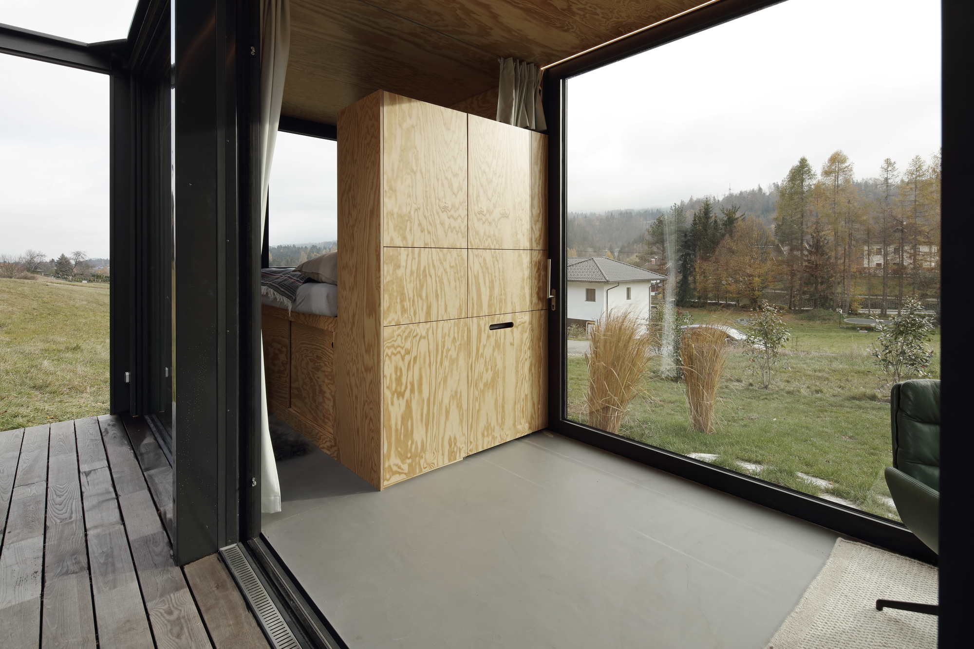 Interior corner with plywood storage wall and floor-to-ceiling glazing opening to autumn landscape
