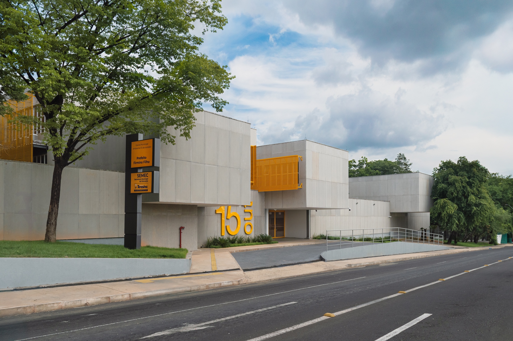 Street facade showing concrete volumes, yellow metal box, and mature trees framing the entrance