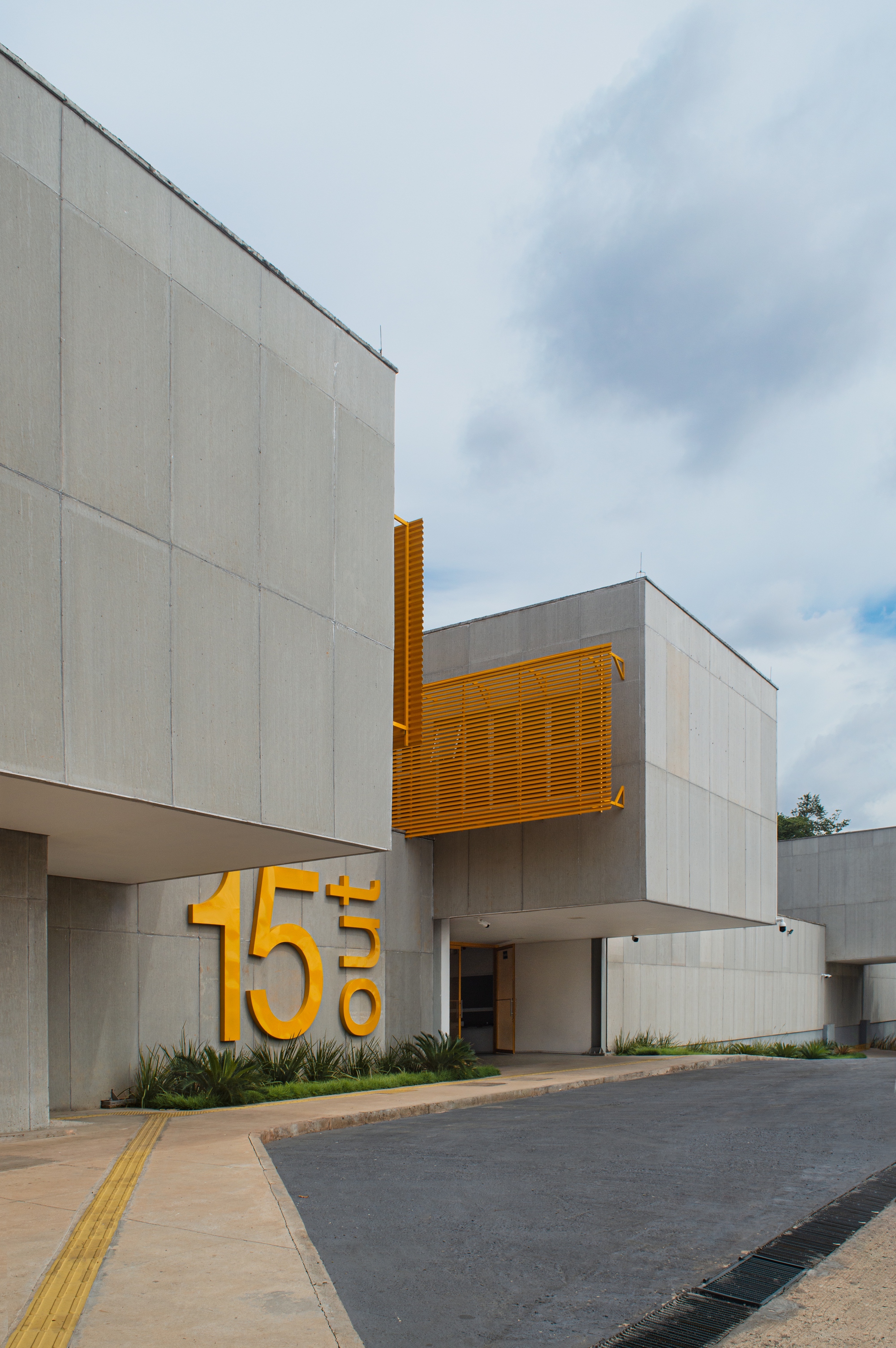 Street entrance with concrete facade, yellow metal louvers, and large dimensional numerals beside planted beds under cloudy sky