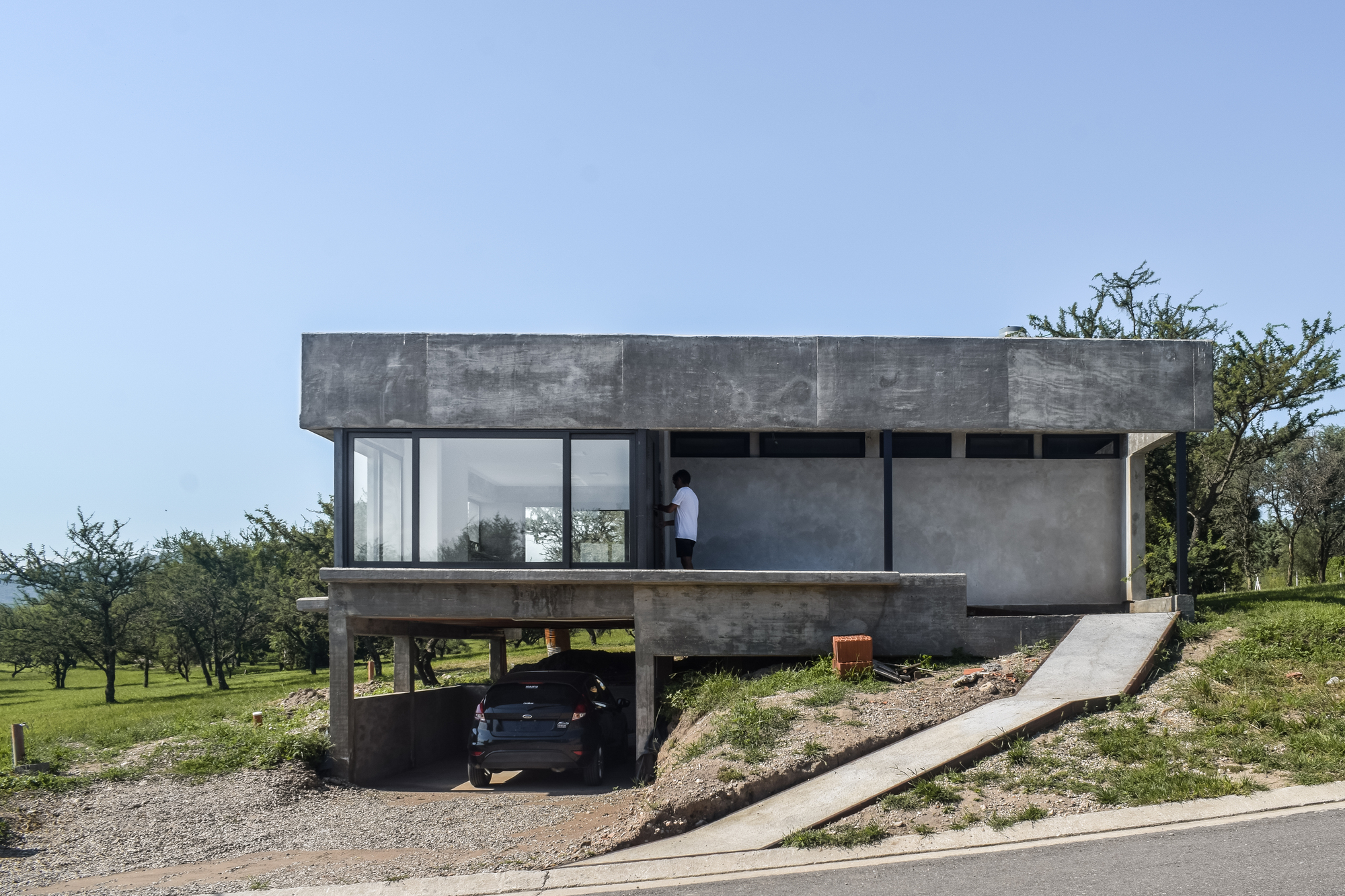 Elevated concrete volume on pilotis with full-height glazing and a figure standing on the terrace