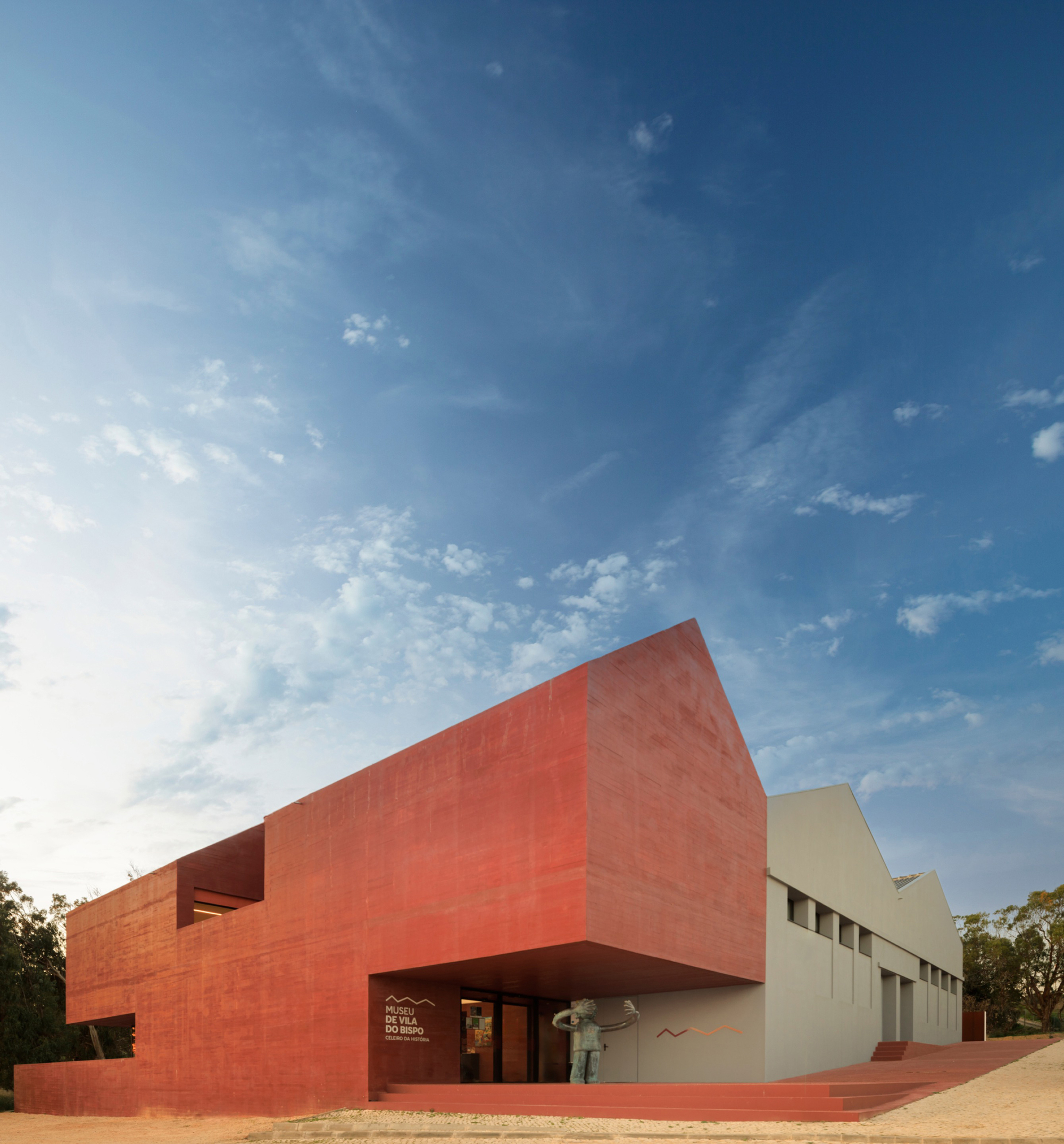Red clay tile facade with cantilevered volume above the entrance against a clear afternoon sky