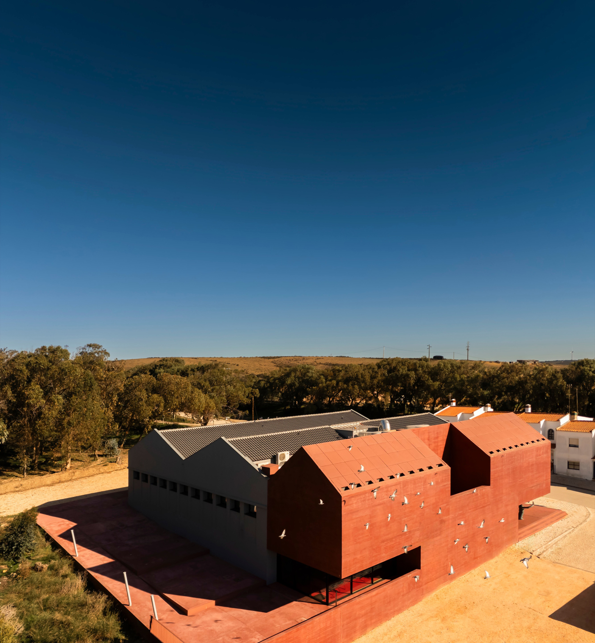 Aerial view of the red tile-clad volumes and metal roofs set in open landscape