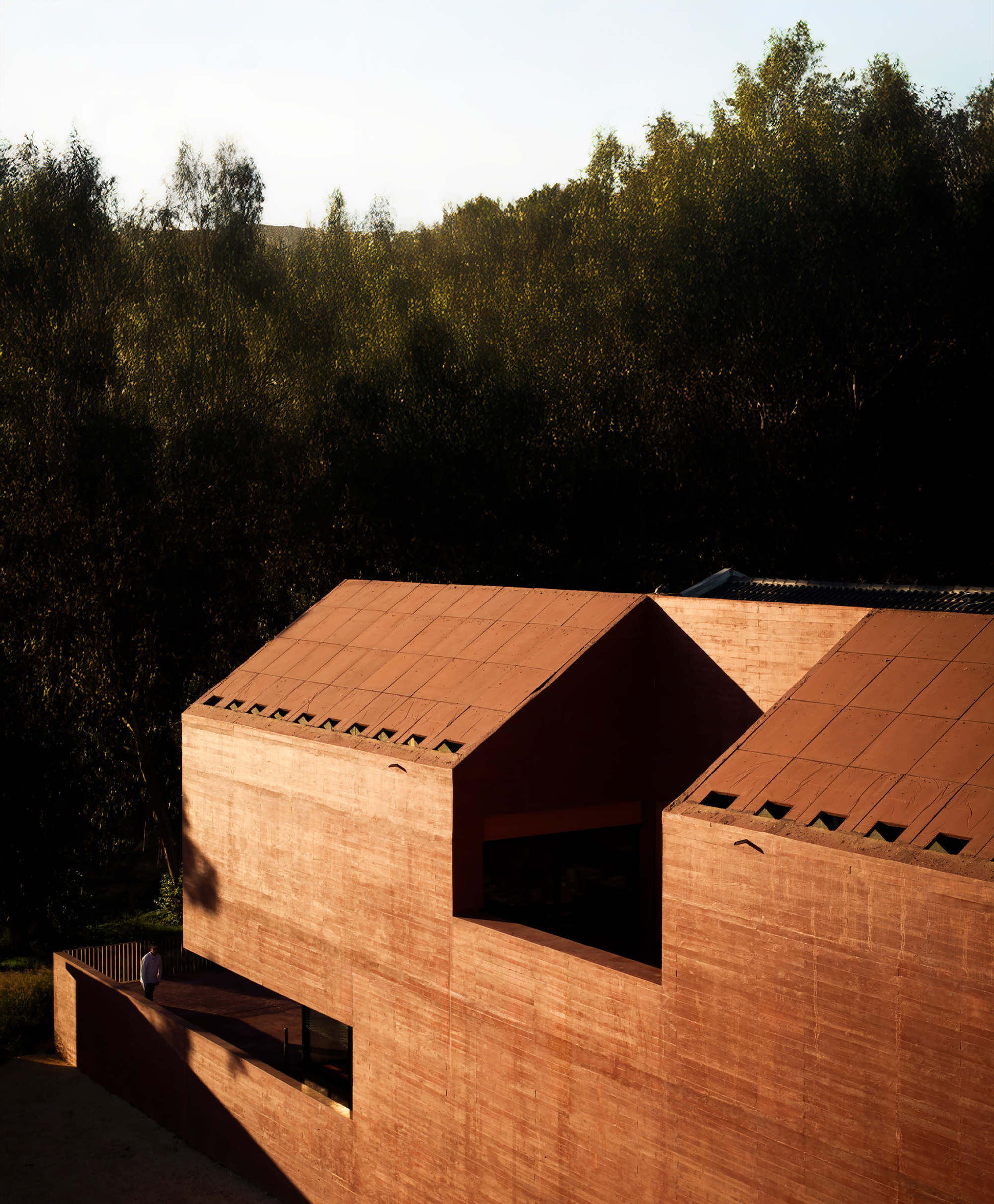Terracotta brick gable volume with deep-set openings in sloped roof at golden hour among trees