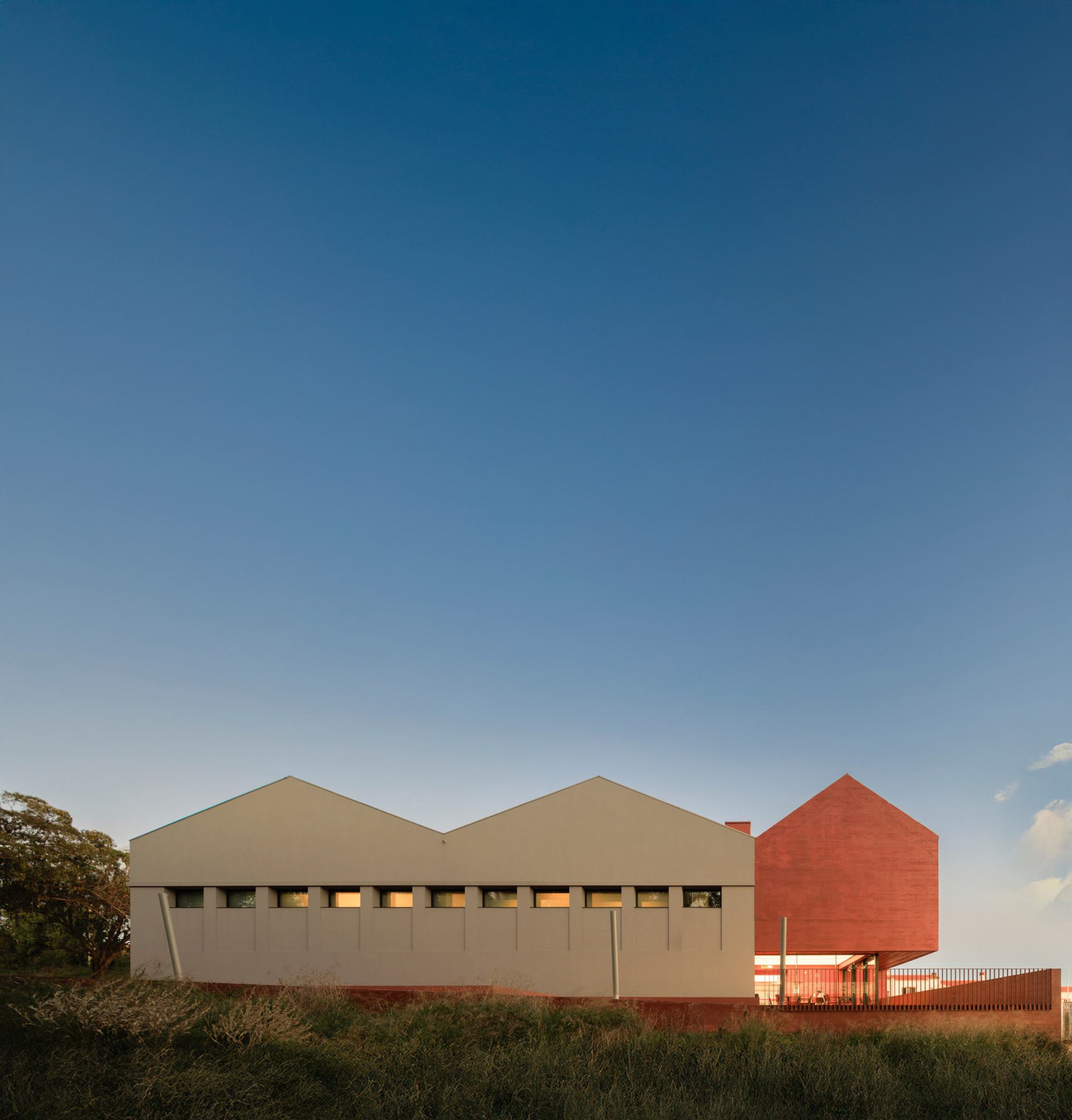 Two gabled volumes clad in beige and red brick alongside a grassy field at dusk