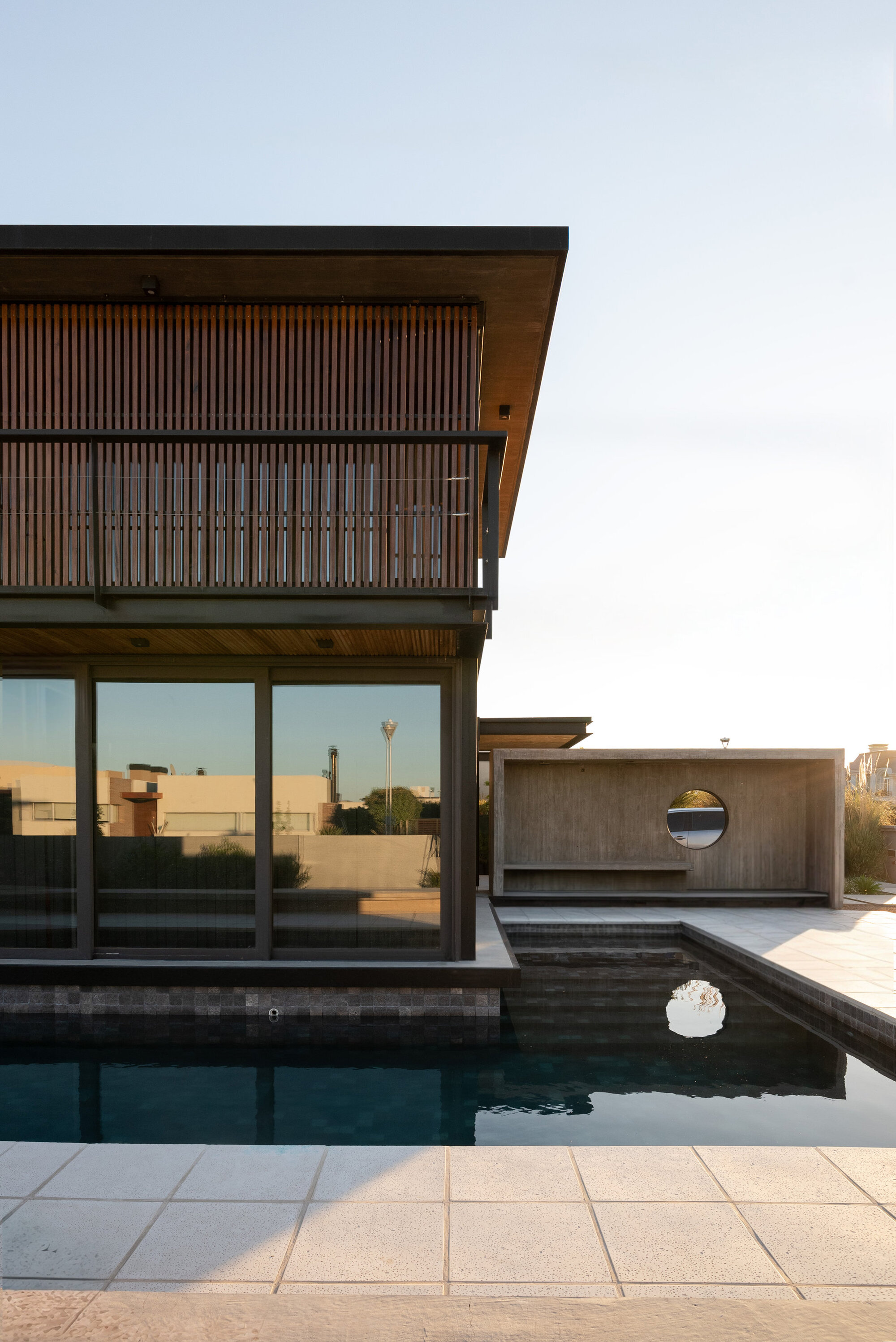 Upper-level terrace with slatted timber screens and cantilever overhang above a pool and circular window volume