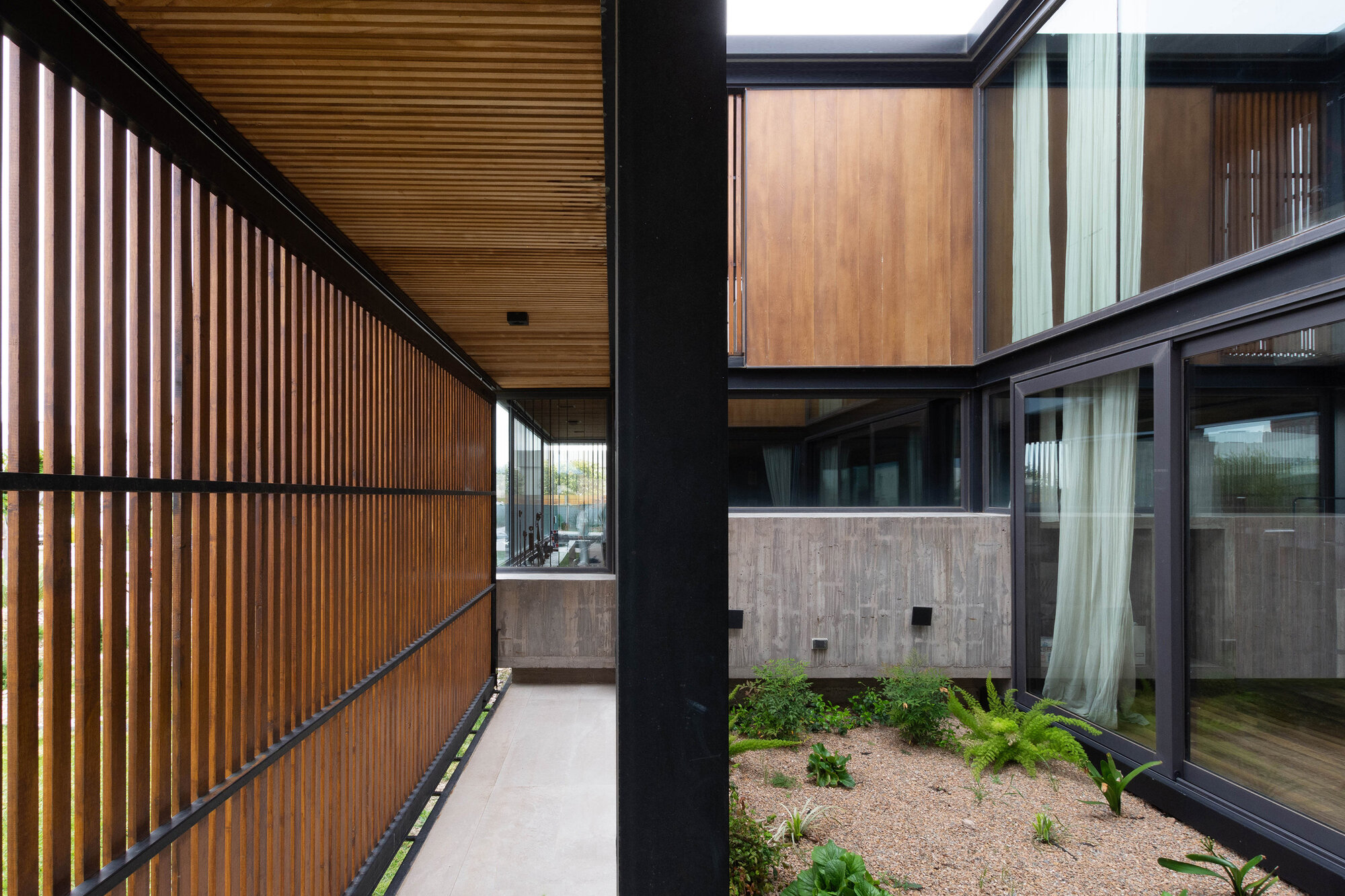 Covered walkway with timber slat screens overlooking a planted courtyard with concrete walls and gravel bed