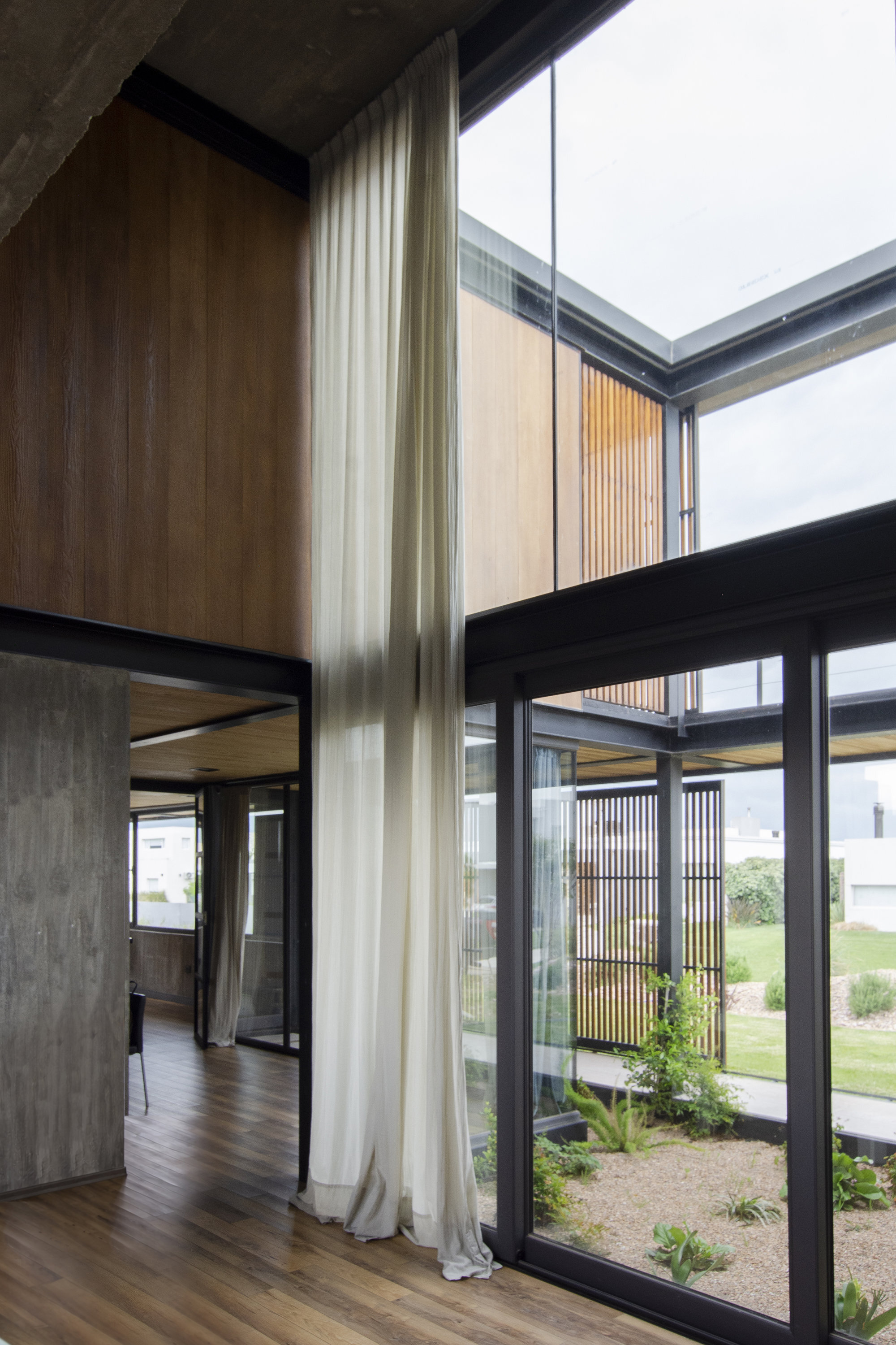 Double-height interior corner with floor-to-ceiling glazing, timber screens, and a planted courtyard visible beyond