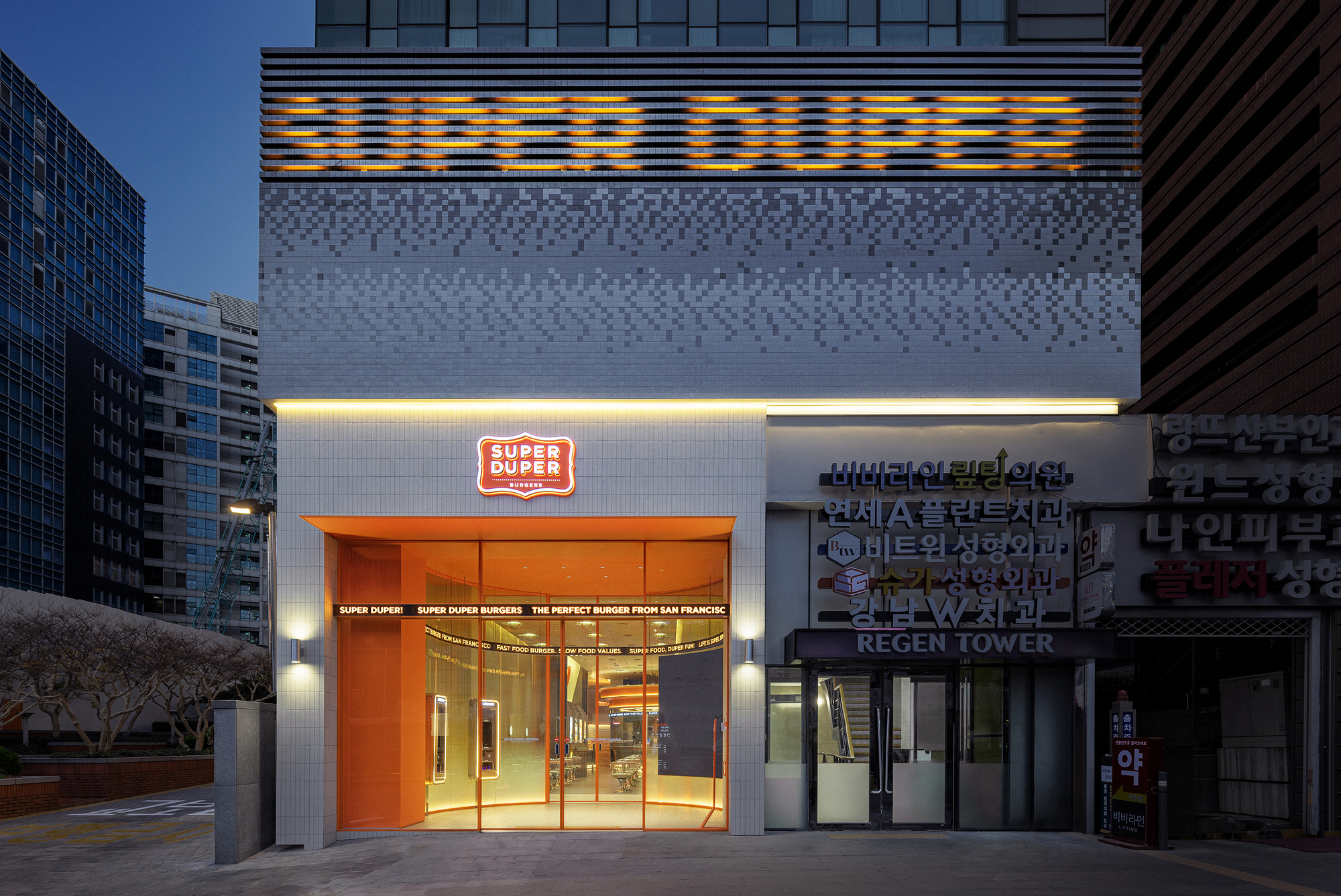 Entrance facade with orange metal doors and illuminated signage at twilight between neighboring buildings