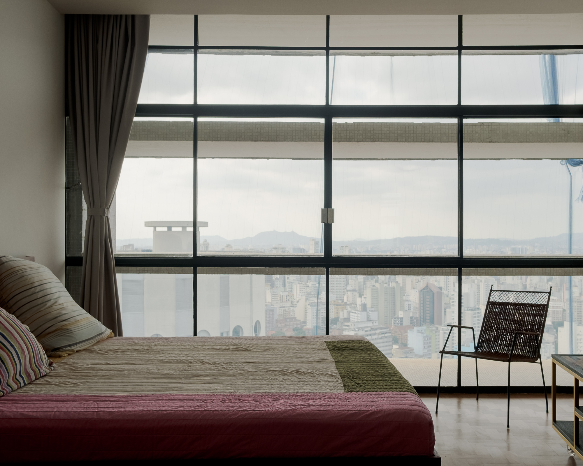 Floor-to-ceiling glazing framing a hazy cityscape view beyond a bed with green runner