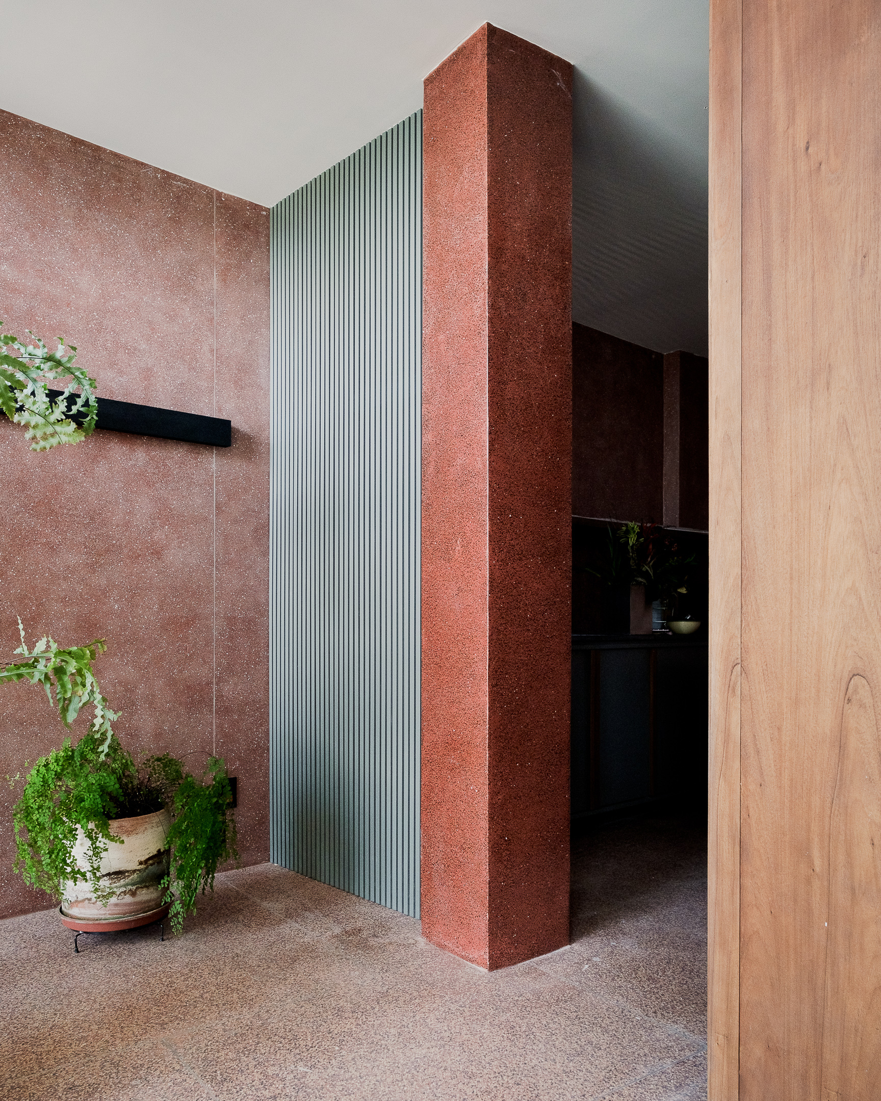 Entrance corridor with red textured stone column, ribbed metal panel and potted ferns on terrazzo floor
