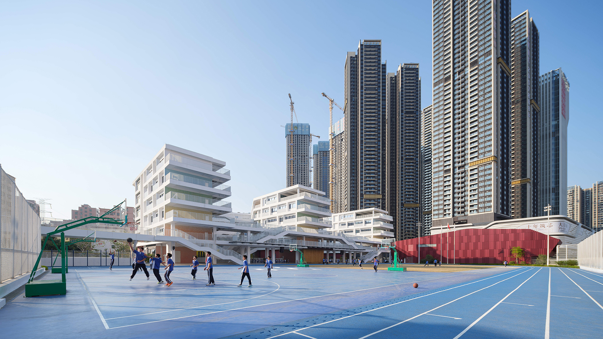 Blue basketball court with students playing in front of white terraced building and highrise towers