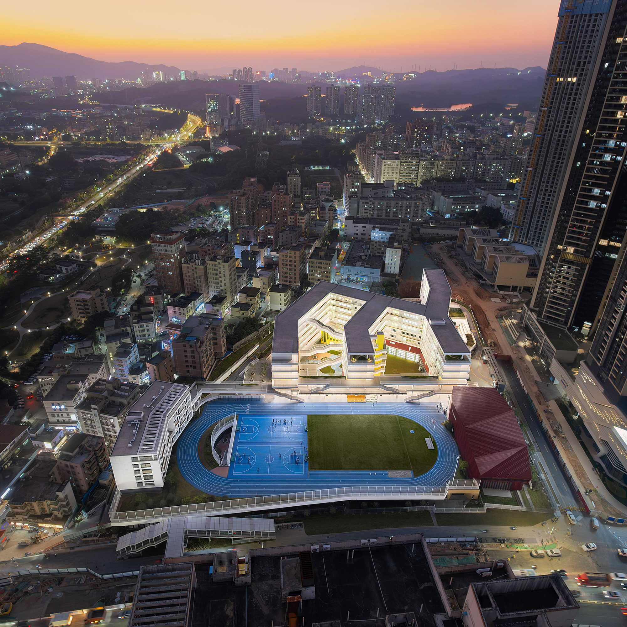 Elevated view of the illuminated white stepped buildings and rooftop sports facilities at sunset with surrounding neighborhood
