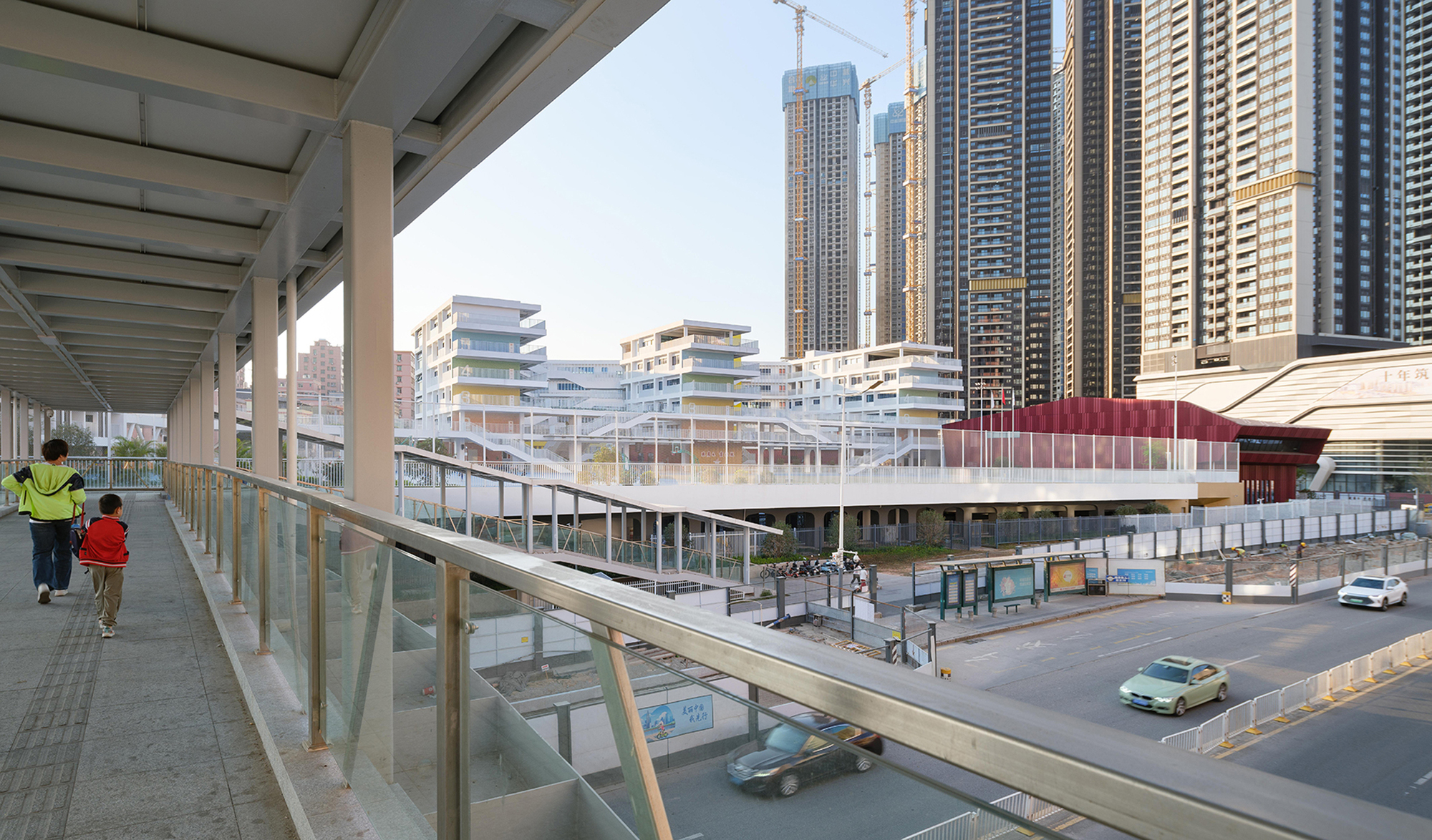 Pedestrian bridge with glazed barrier overlooking the layered white school complex and street below in daylight
