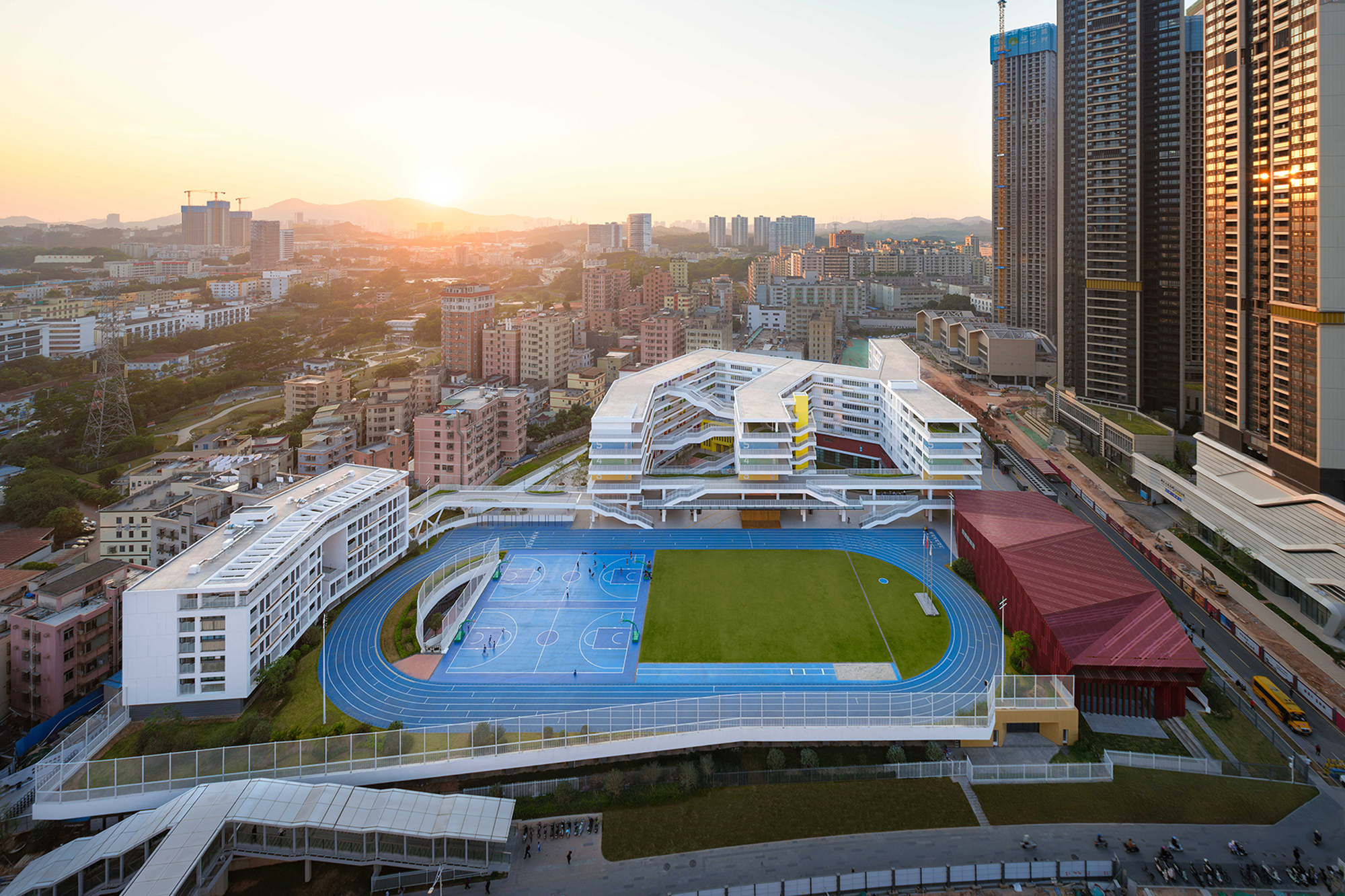 Oblique aerial view of the white cascading school volumes and rooftop athletic facilities at sunrise with mountains beyond
