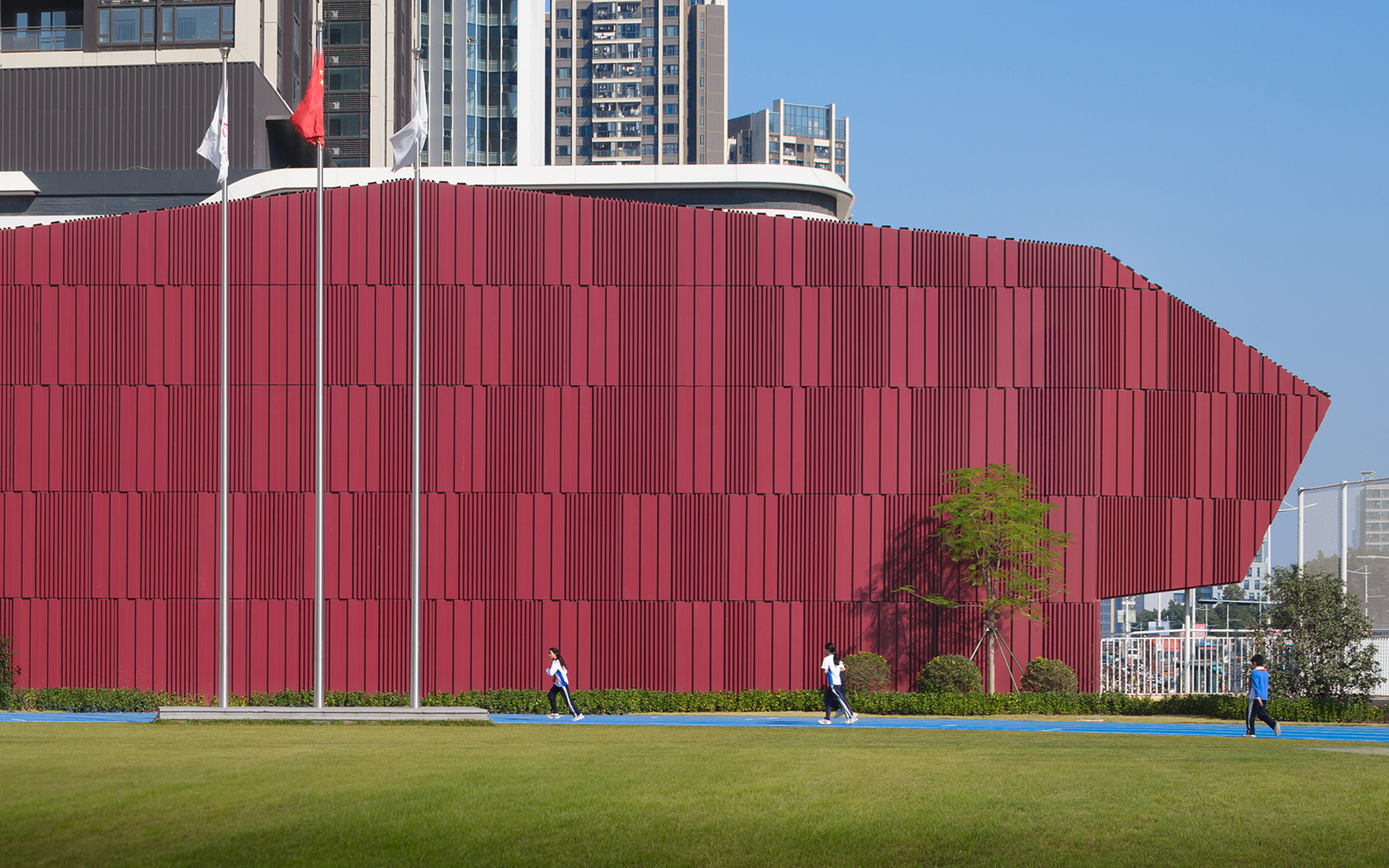 Red vertically ribbed facade with flags above, adjacent to a green lawn with pedestrians and trees