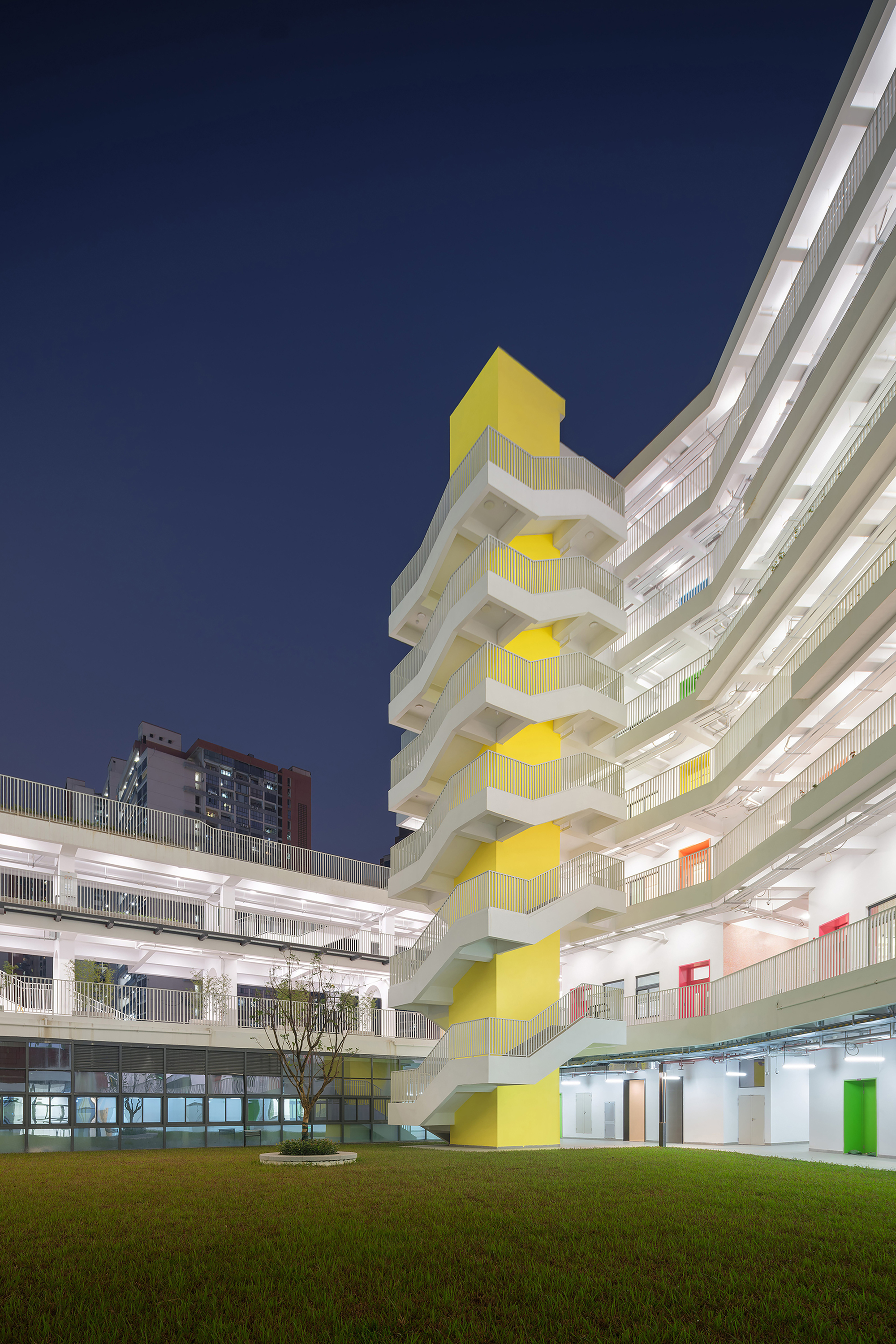 Yellow stacked staircase tower illuminated against a dark blue evening sky beside white terraced school buildings